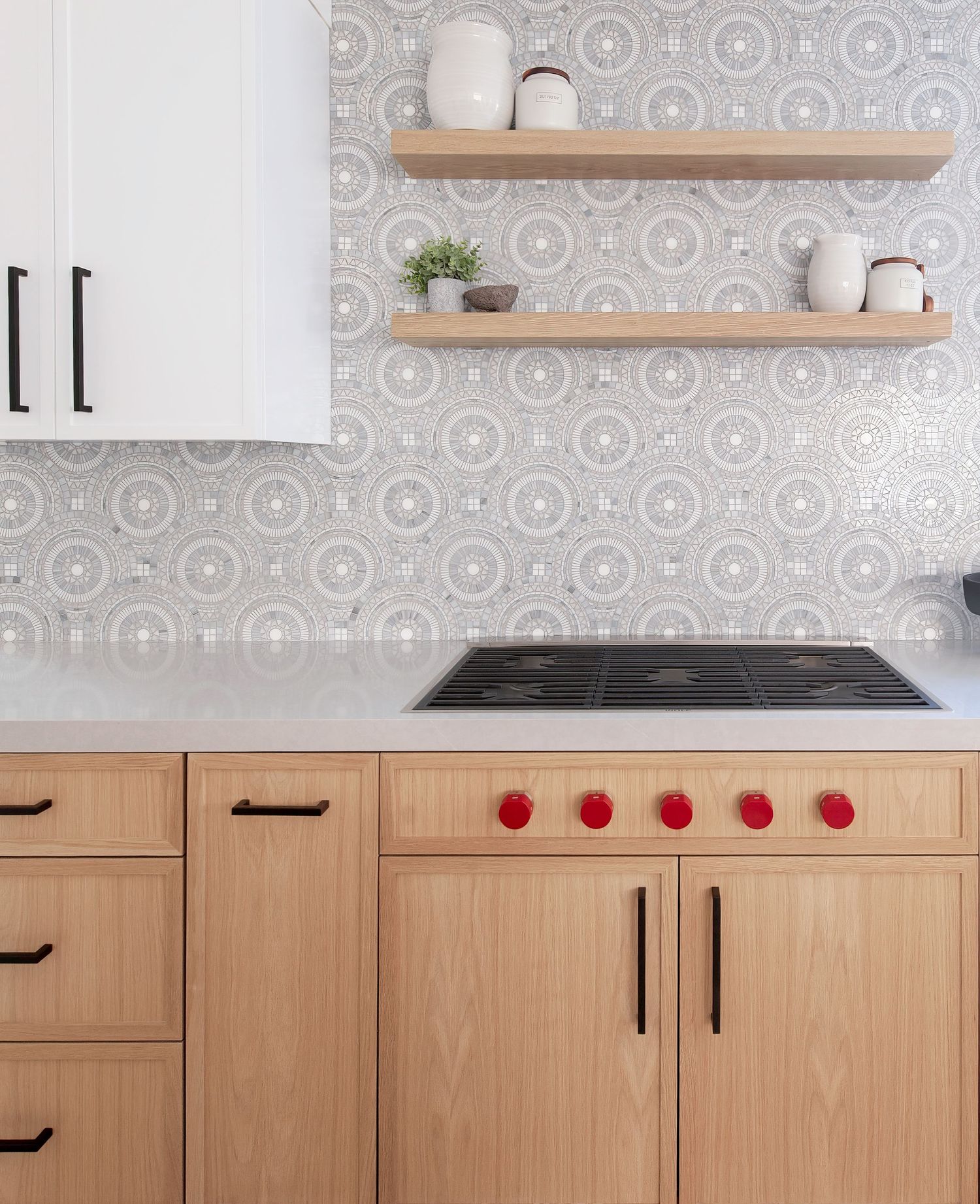 Modern kitchen featuring light wood cabinets and geometric patterned backsplash with red knobs.