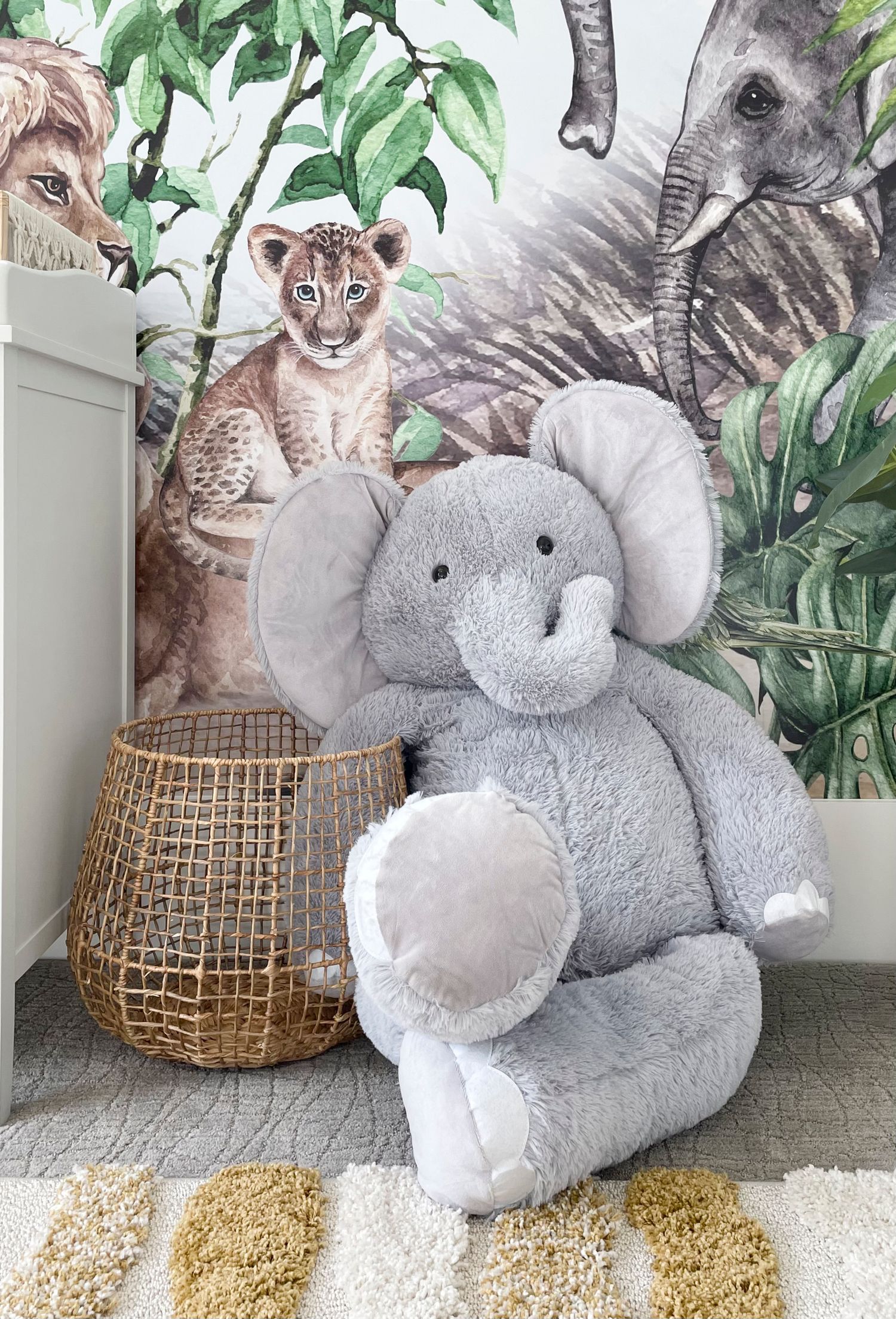 Nursery vignette of a large gray plush elephant toy sitting beside a woven basket against a tropical leaf wallpaper background.