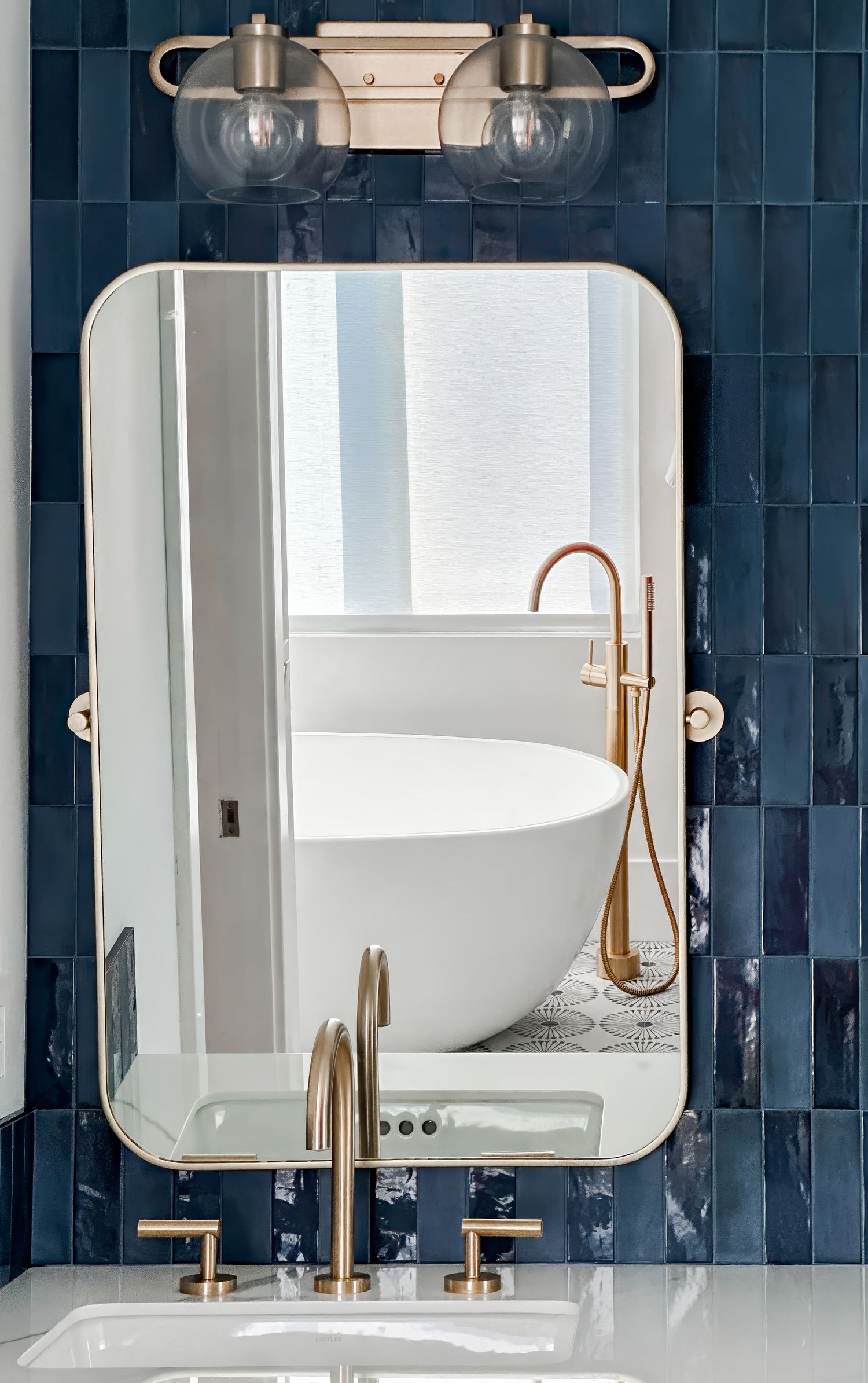Creative photo of a modern designed bathroom featuring navy blue tile walls, rounded mirror, and brass fixtures.