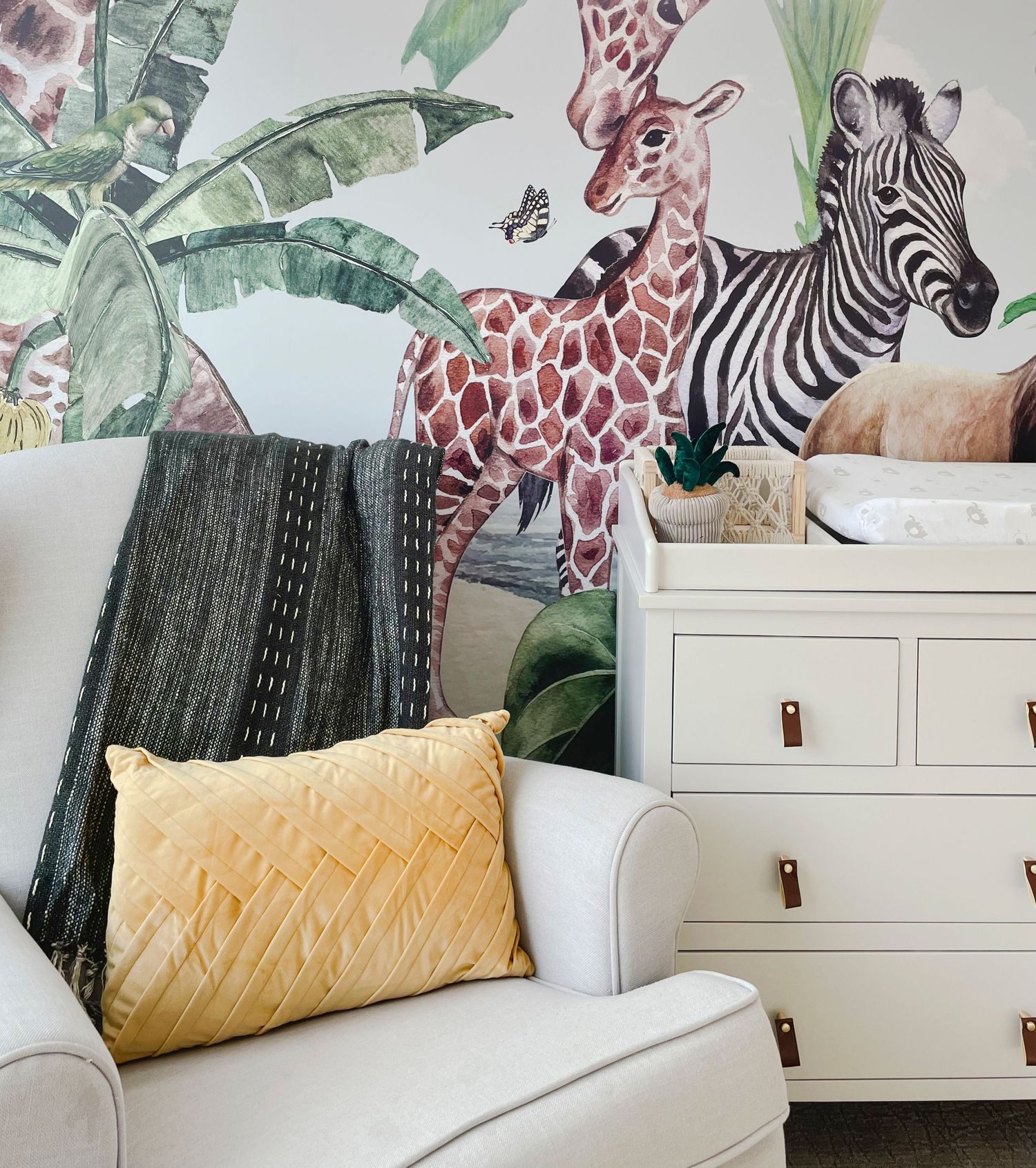 Nursery corner featuring whimsical safari animal wallpaper, white furniture, and yellow accent pillow.