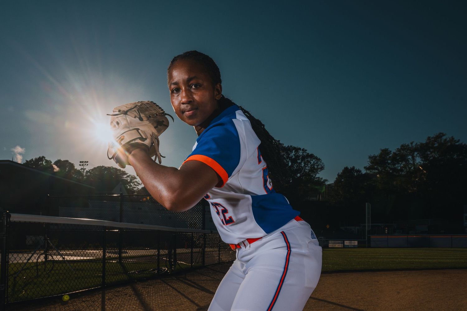 A baseball pitcher demonstrates throwing stance against dramatic backlit sky.