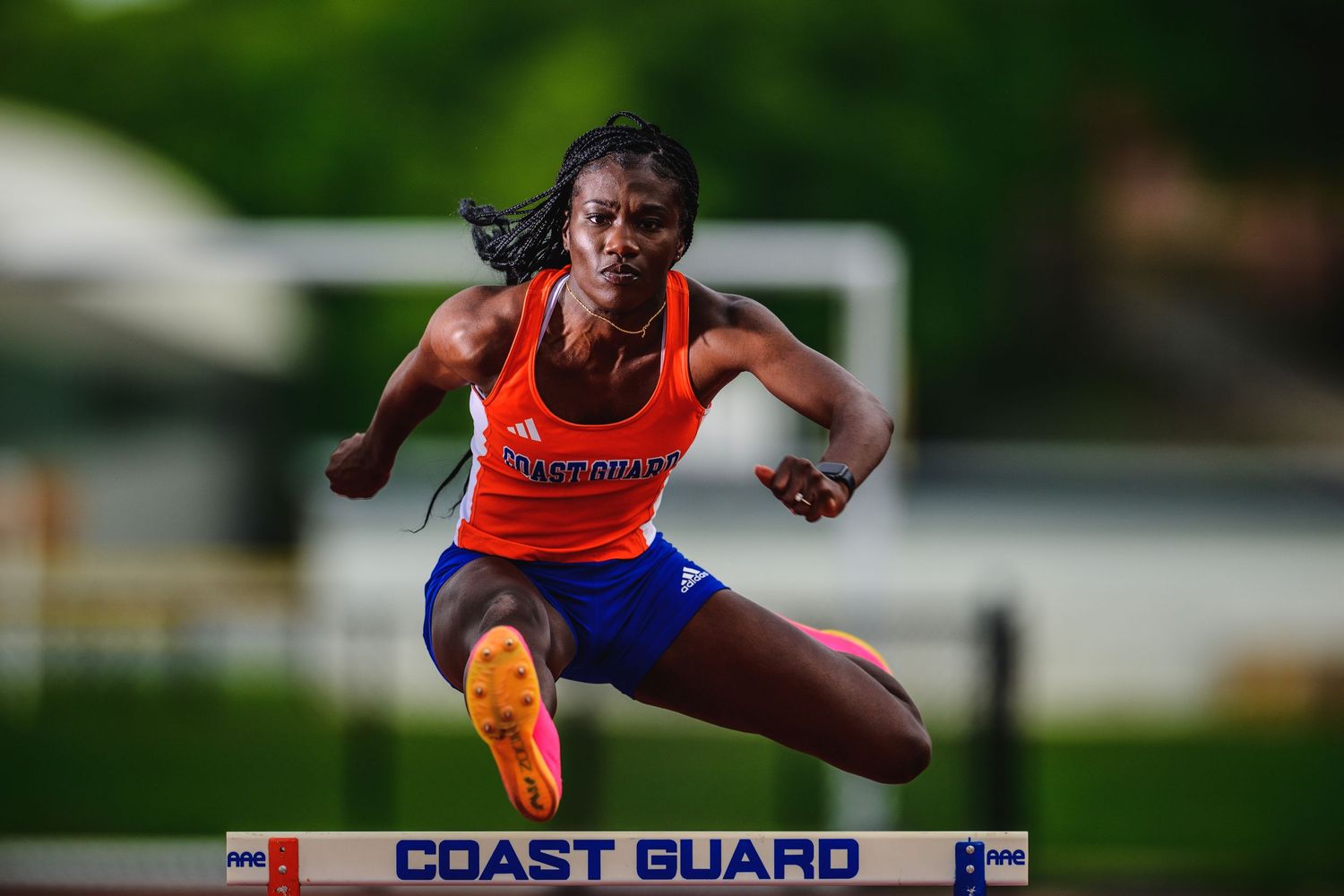Hurdler in orange uniform clears obstacle during Coast Guard track meet.