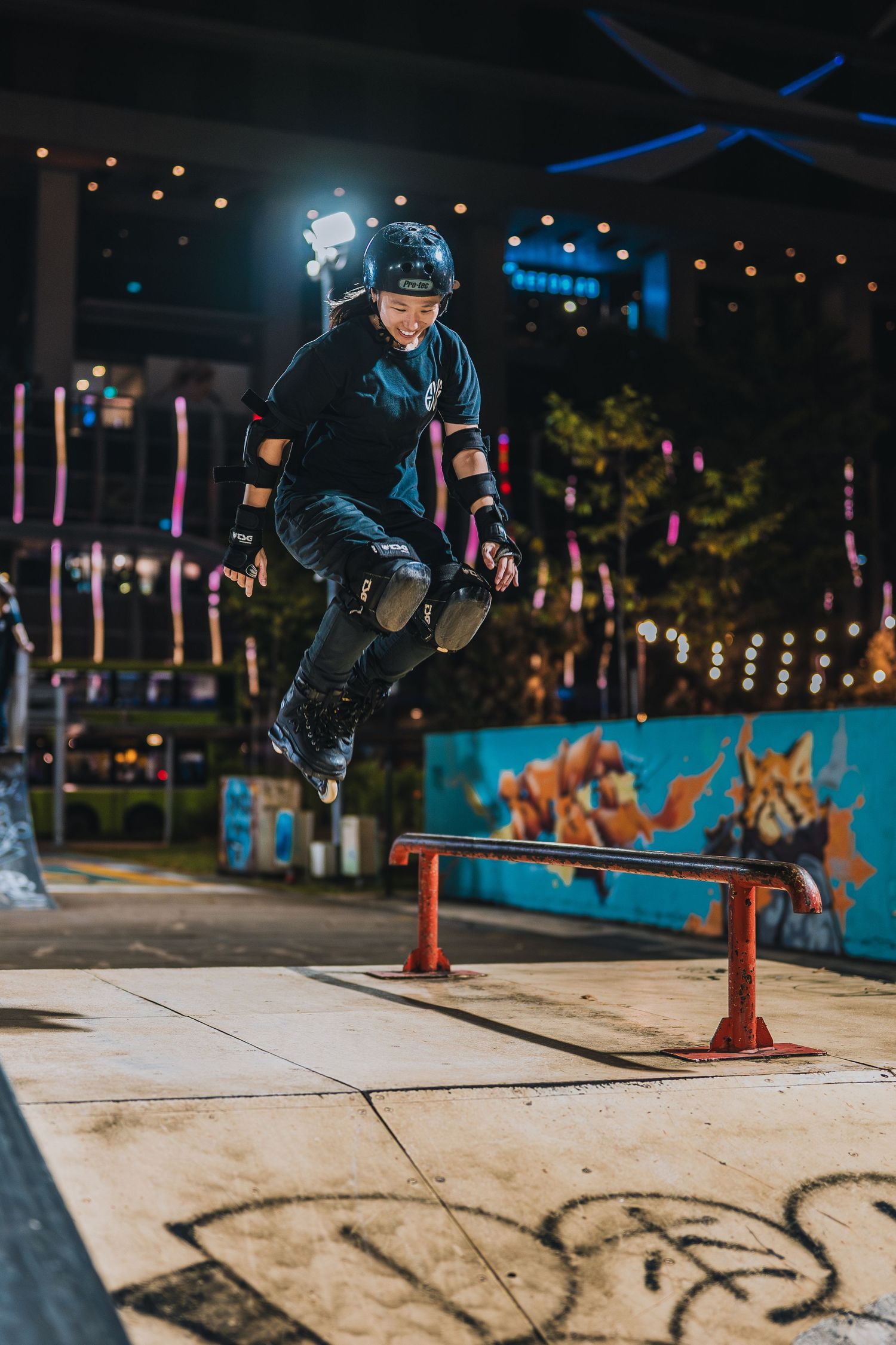 Rollerblader jumping over metal rail at illuminated urban skate park with colorful mural backdrop.