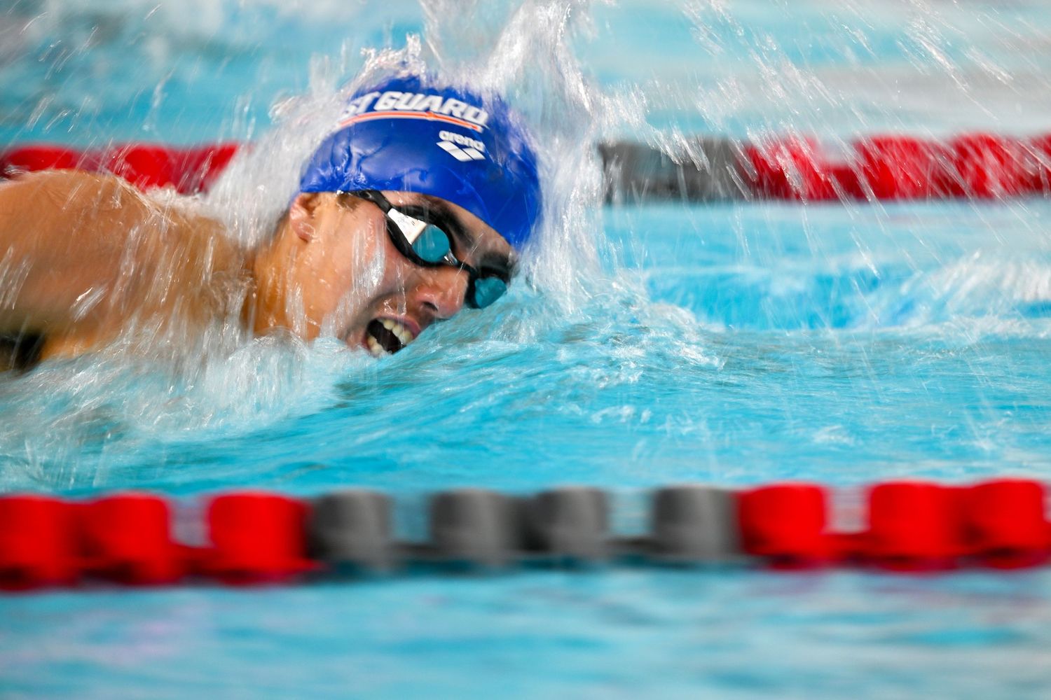 A swimmer in a blue cap performs the butterfly stroke during a competitive race in a pool with red lane markers.