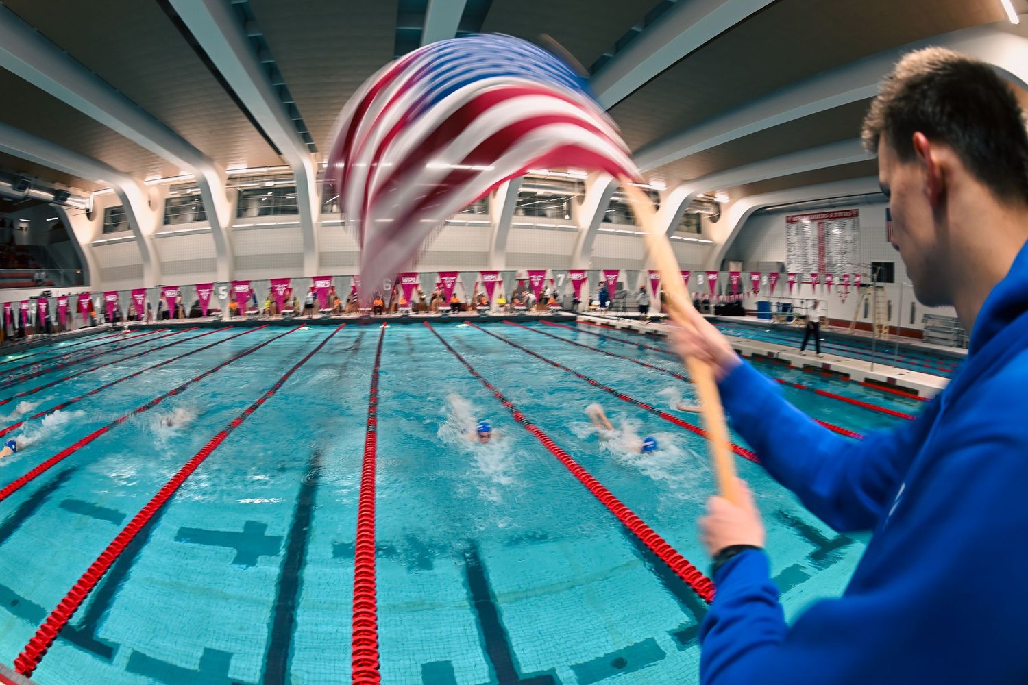 American flag waving over indoor competitive swimming pool with lane dividers at aquatic center competition.