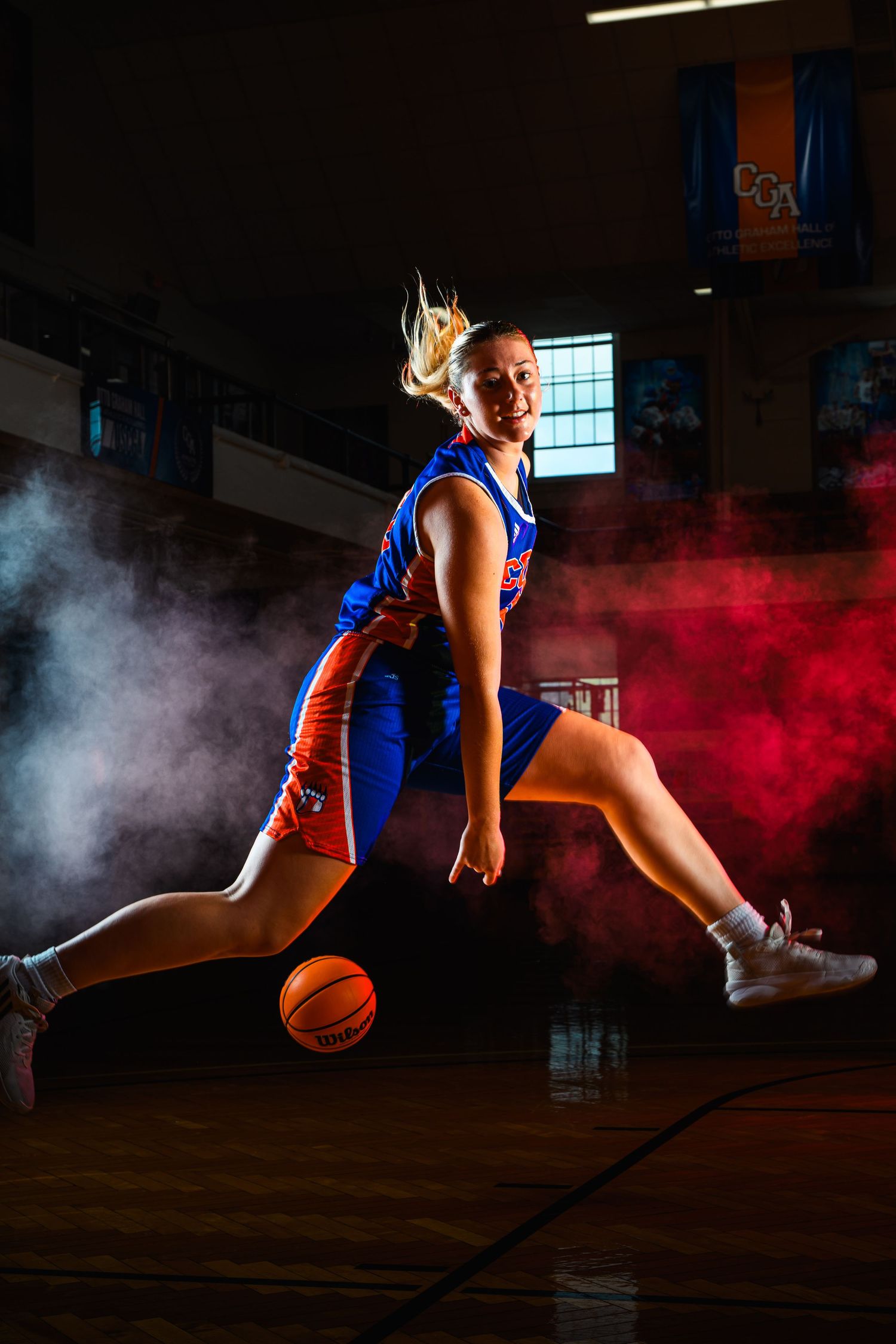 Dramatic sports photo of an athlete in blue uniform dribbling a basketball through colored smoke effects in a gym.