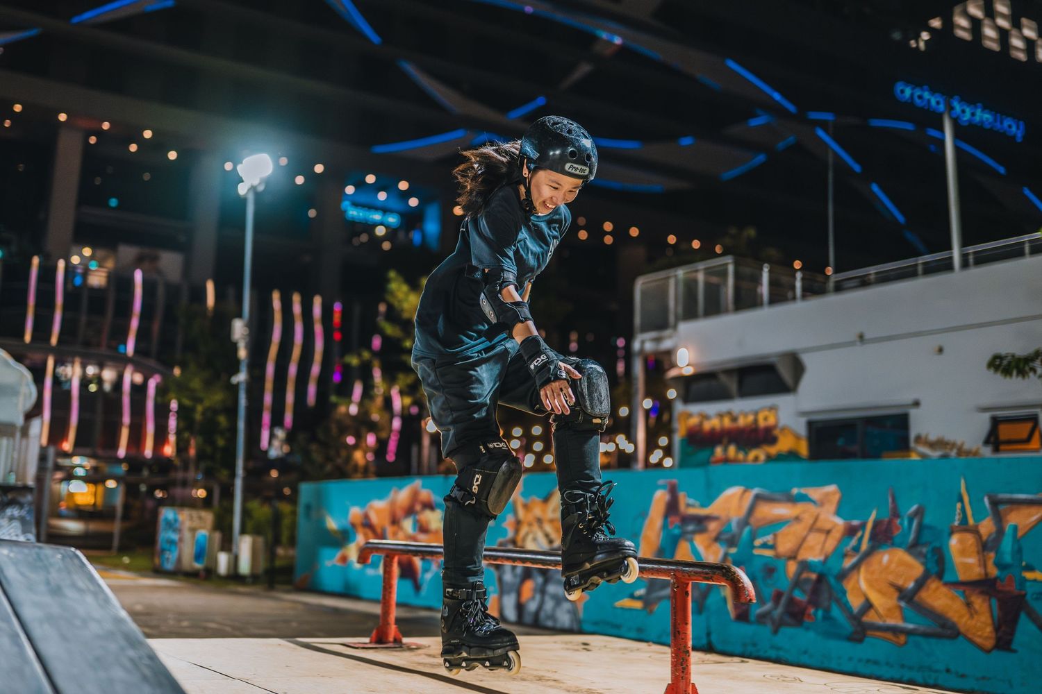Rollerblader performing aerial trick over obstacle with colorful mural and night lights.