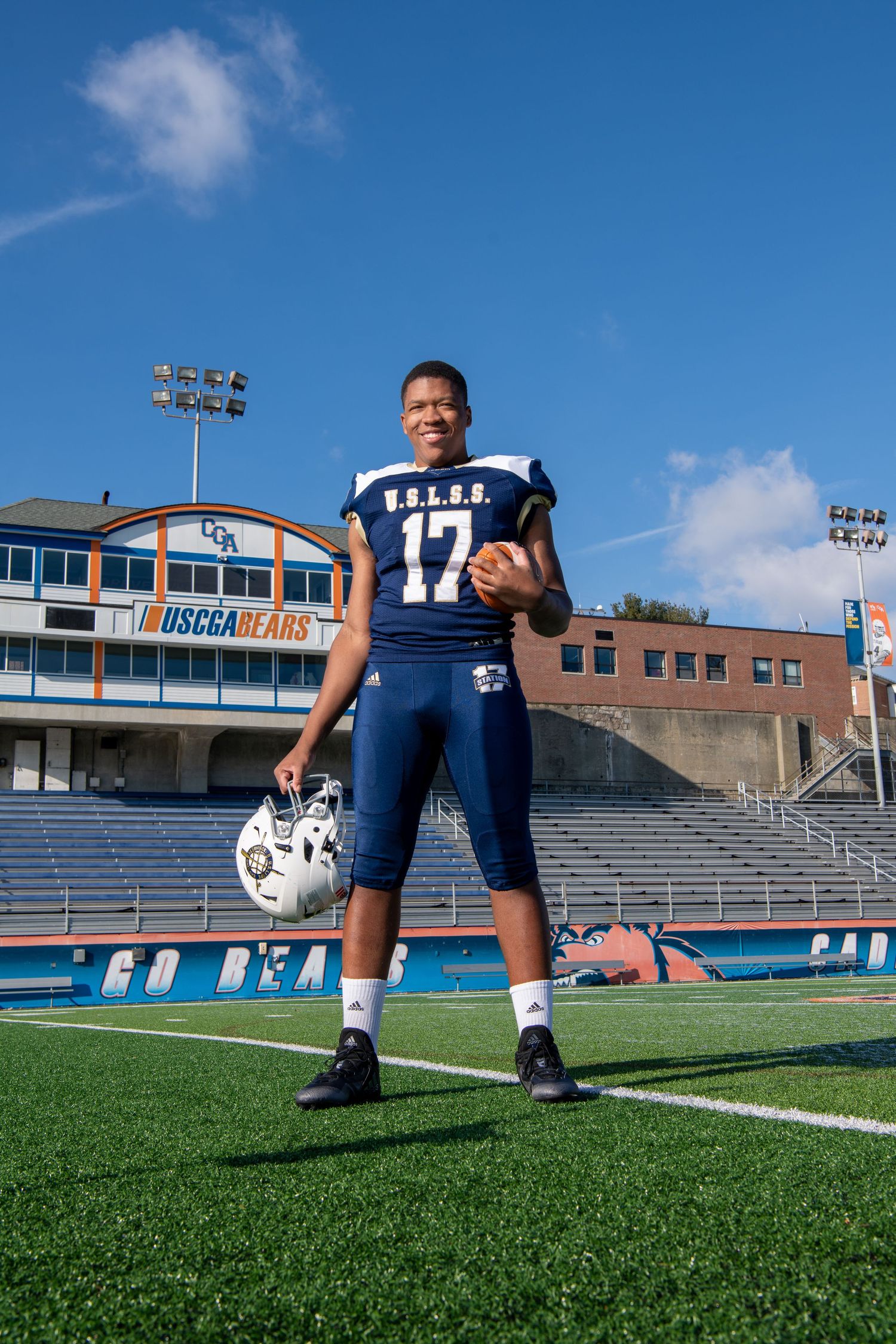 Football player in navy uniform stands on field with stadium in background.