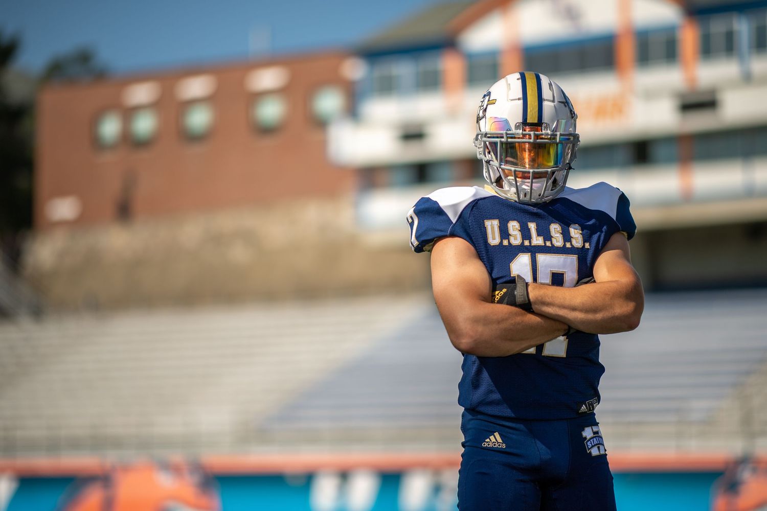 Football player in navy blue jersey and helmet standing on field with crossed arms.
