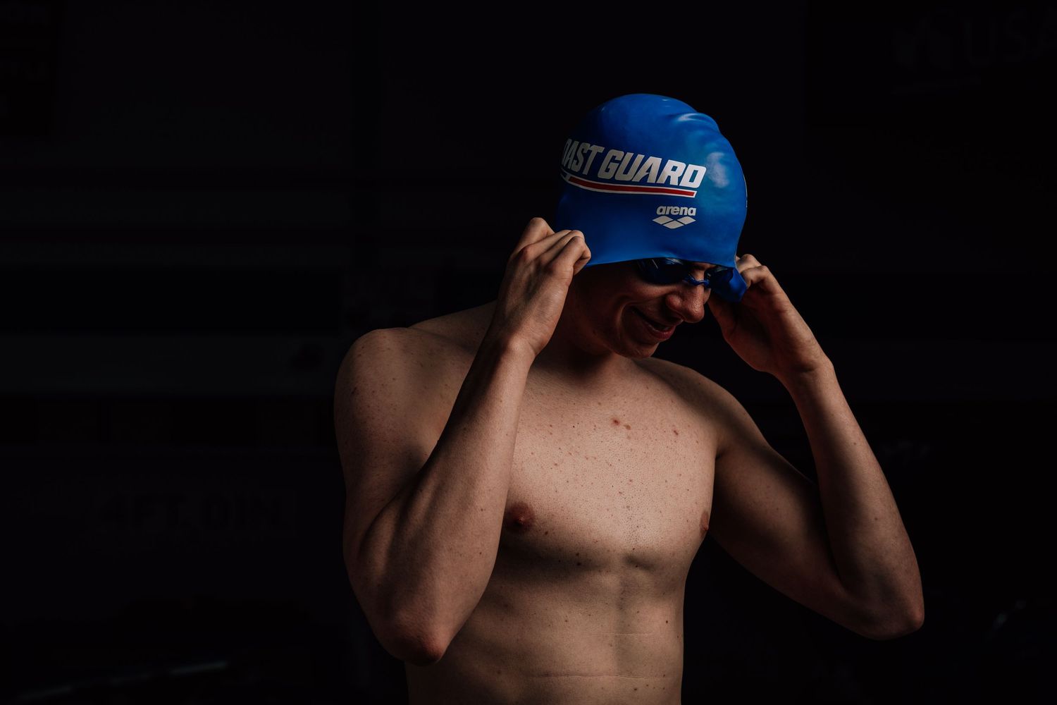 Shirtless athlete adjusting blue swimming cap against dark background in dramatic lighting.