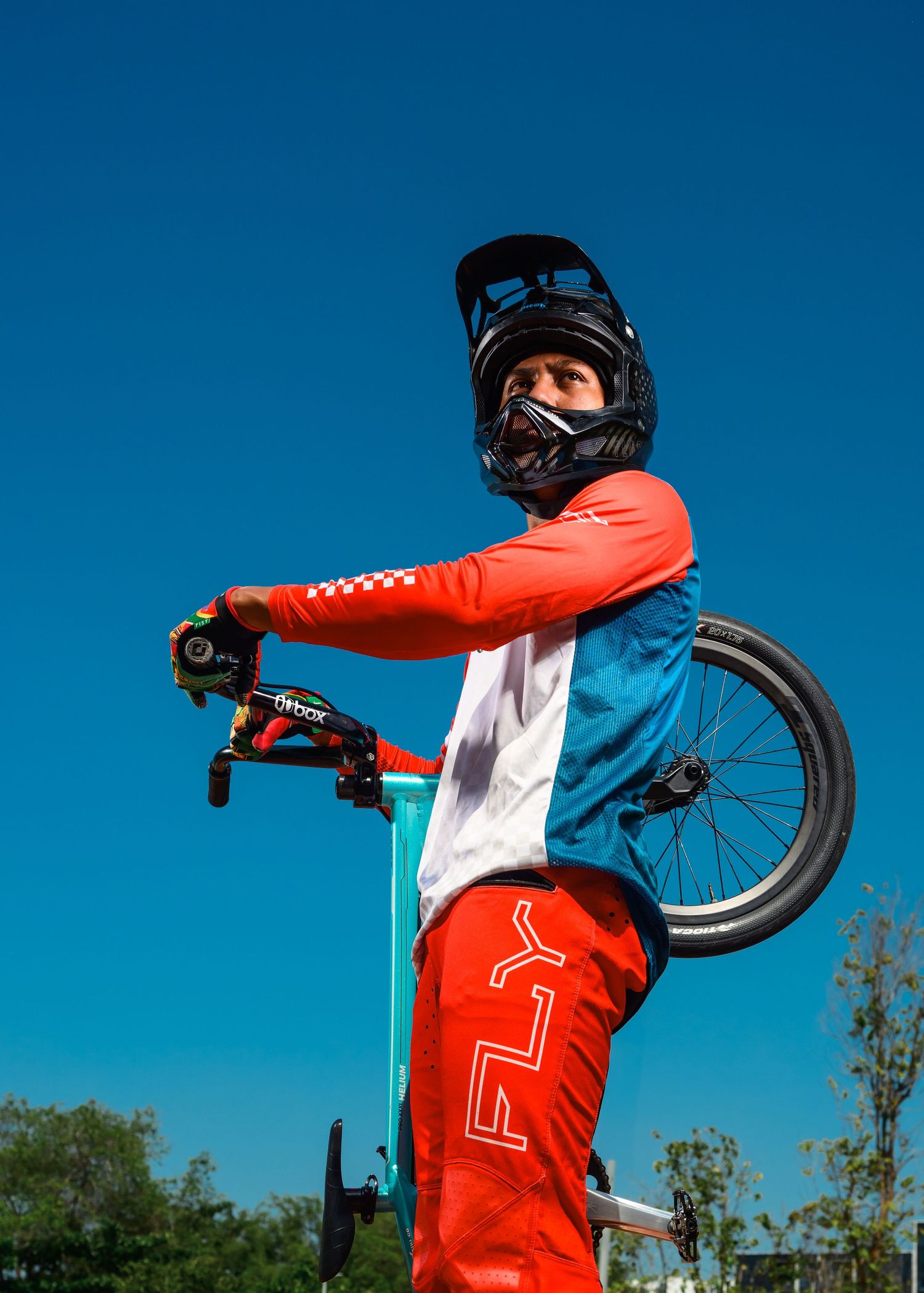 BMX rider in red gear and full-face helmet poses with bike against blue sky.