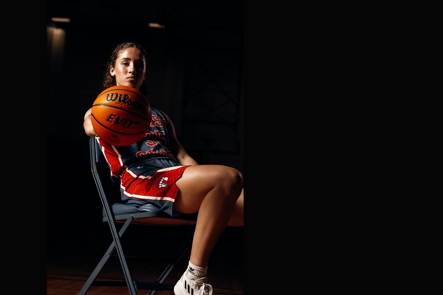 Basketball player sits on blue chair in dramatic studio lighting against dark background.