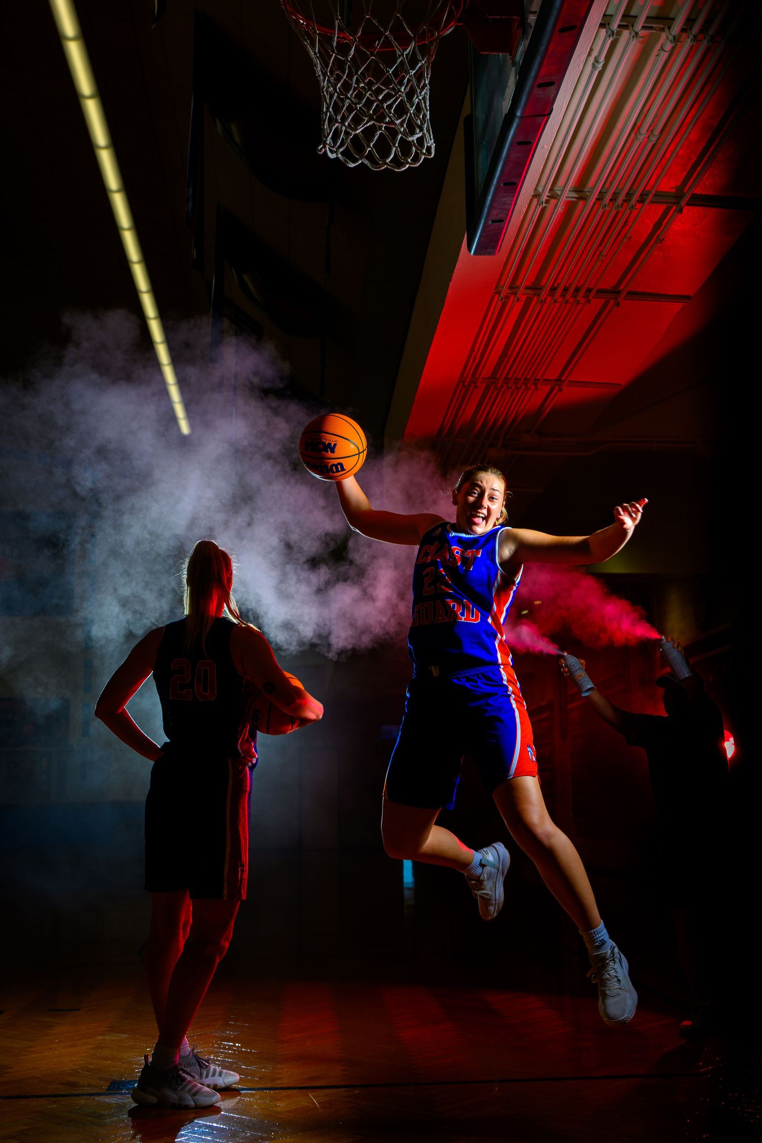 Dynamic basketball action shot showing players jumping towards hoop with dramatic lighting and smoke effects.