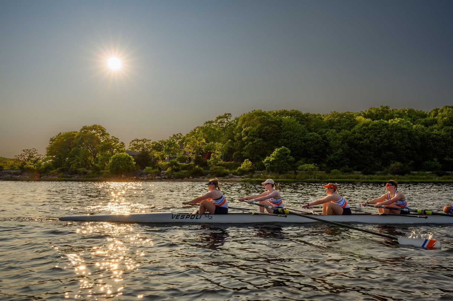 Four person rowing team gliding across water at sunset with sunlight reflection.