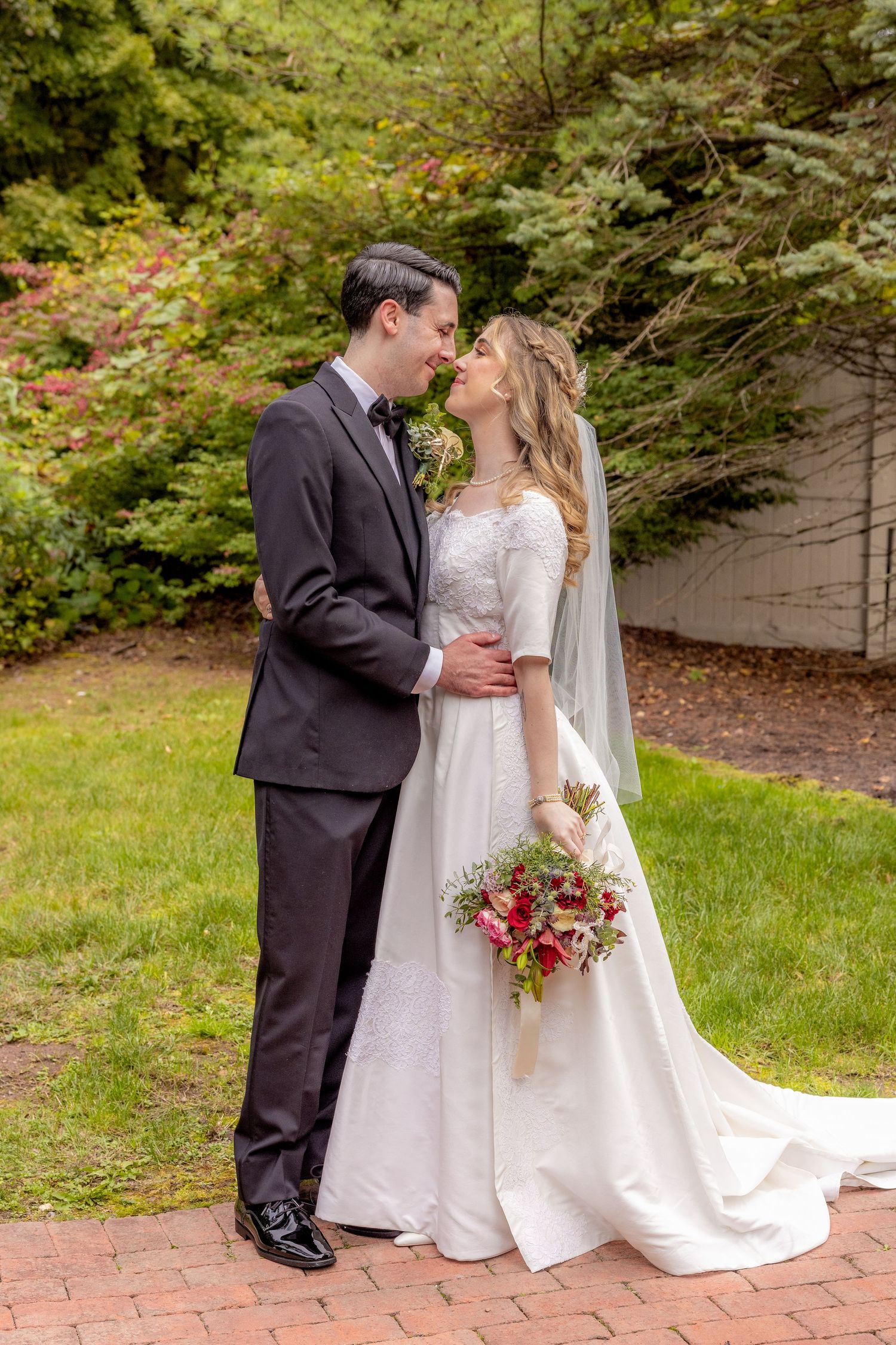 Romantic wedding couple embraces on brick pathway surrounded by lush autumn foliage.