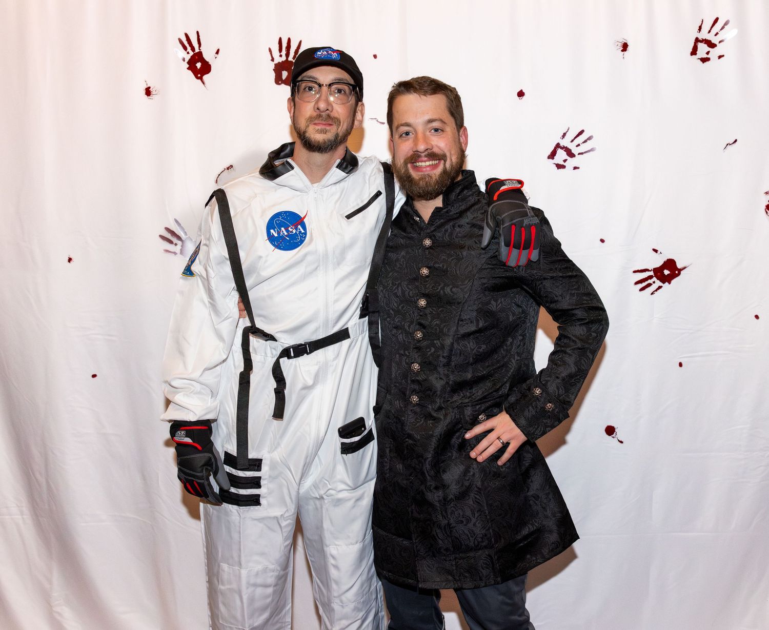 Two people pose in contrasting astronaut and vintage military costumes against a white backdrop with red handprints.