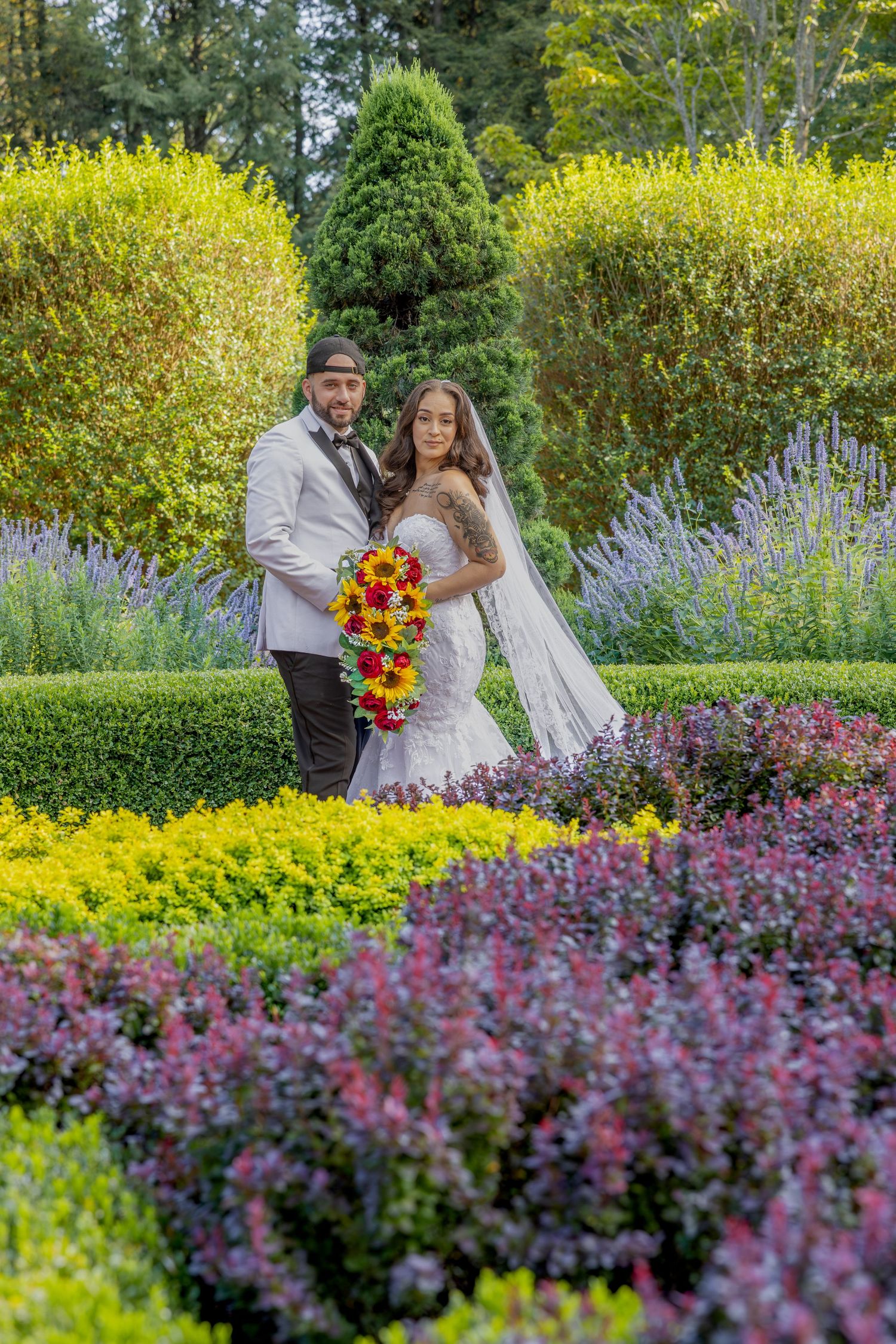 Couple poses in vibrant garden with yellow and purple flowers, bride holding cascading sunflower bouquet.