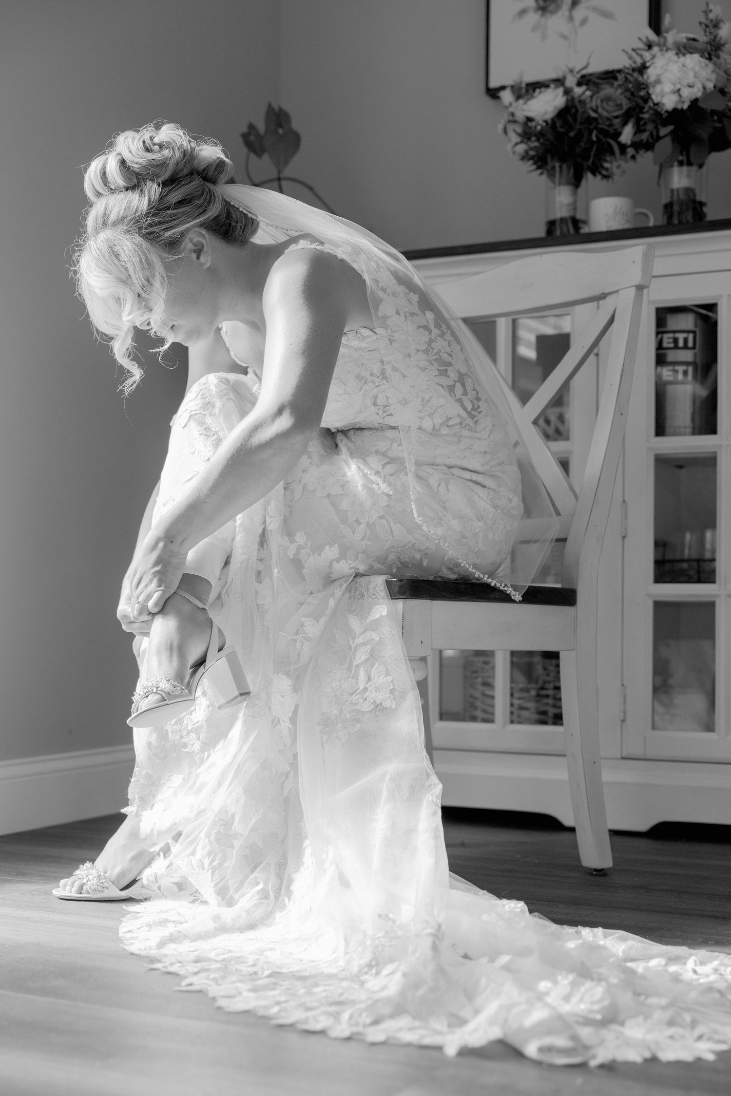 Black and white photo of bride in lace dress adjusting her shoes.