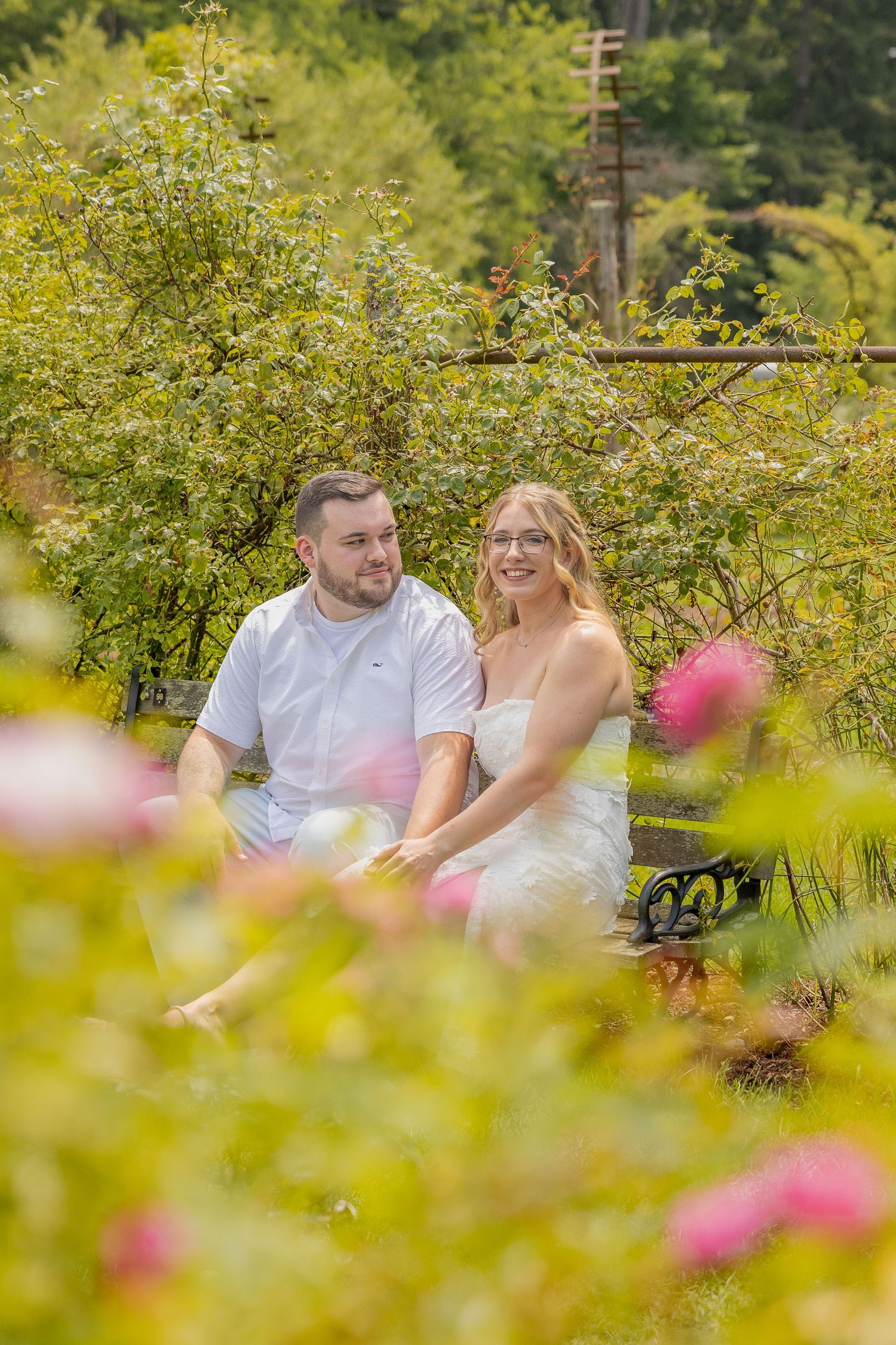 A couple sits together in a lush garden filled with blooming flowers and greenery on a sunny day.