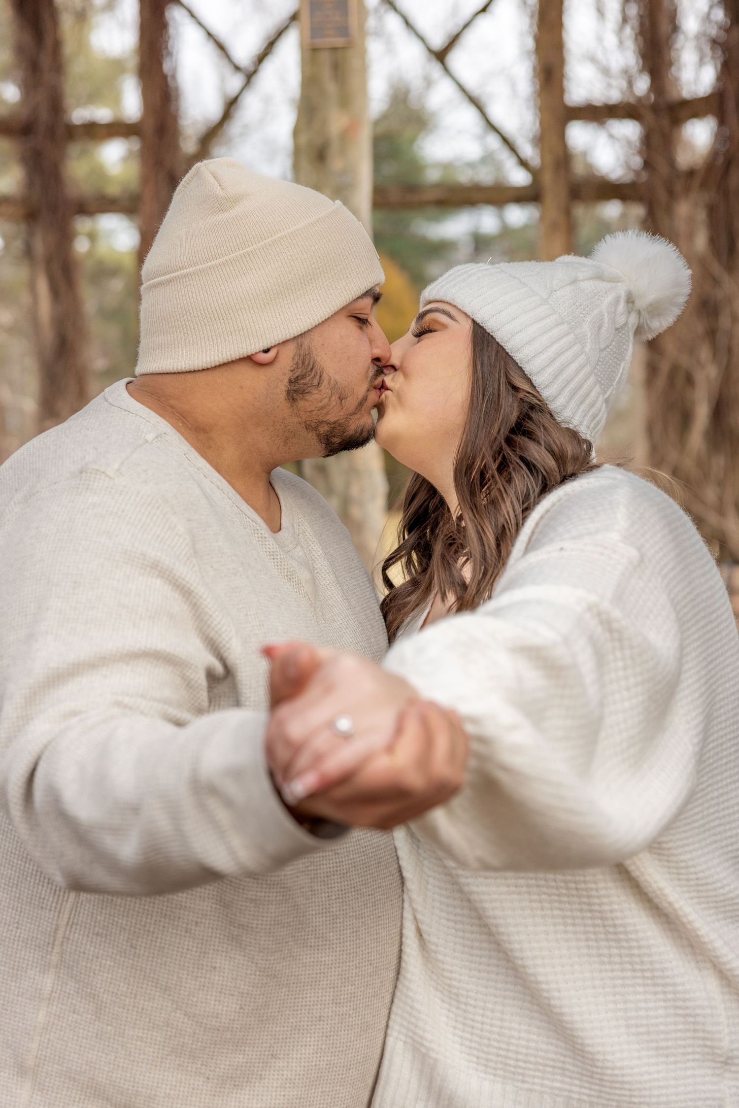 A couple in matching white winter outfits and beanies share a romantic kiss in a wooded setting.