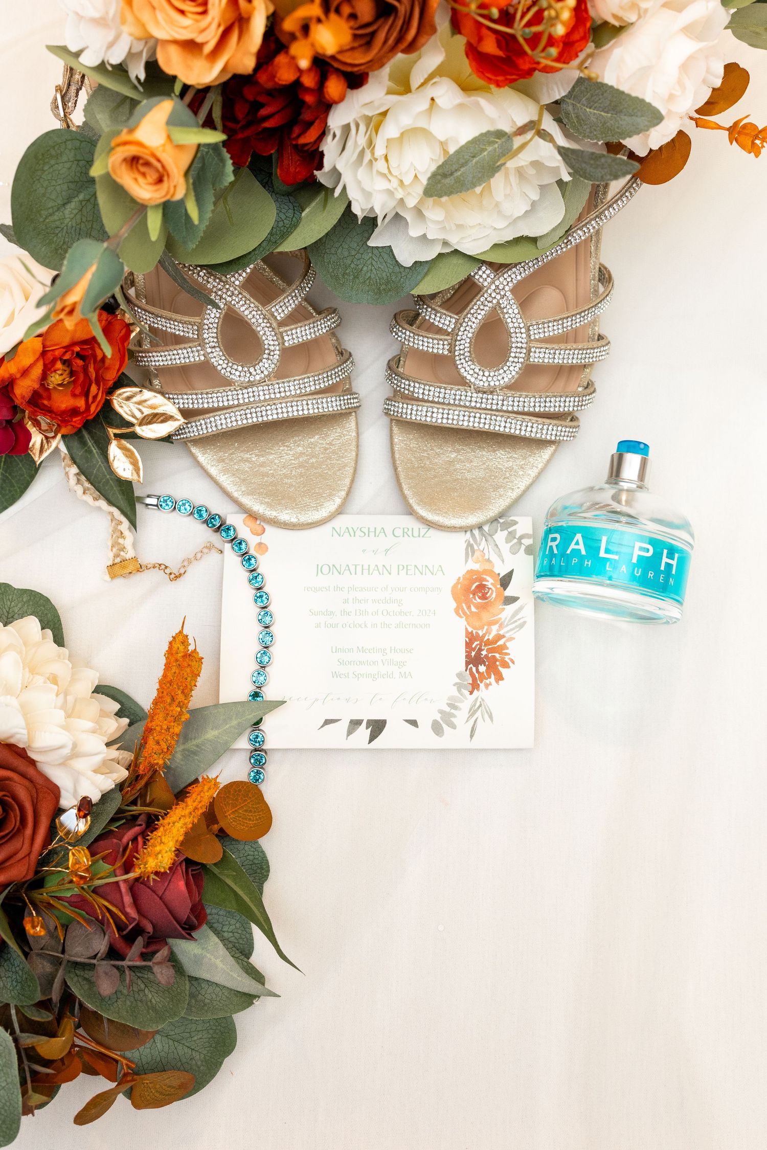Wedding details flatlay with floral arrangements, sparkly sandals and an invitation on white background.