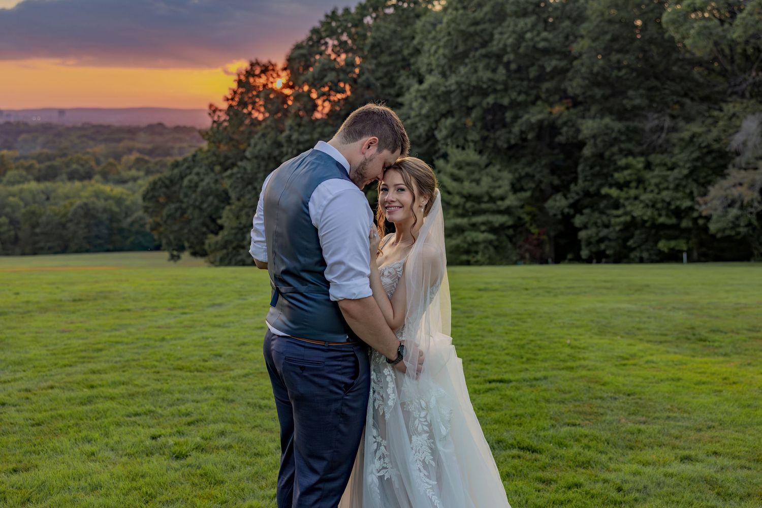 A romantic sunset wedding portrait on a hilltop with golden light filtering through trees in the background.