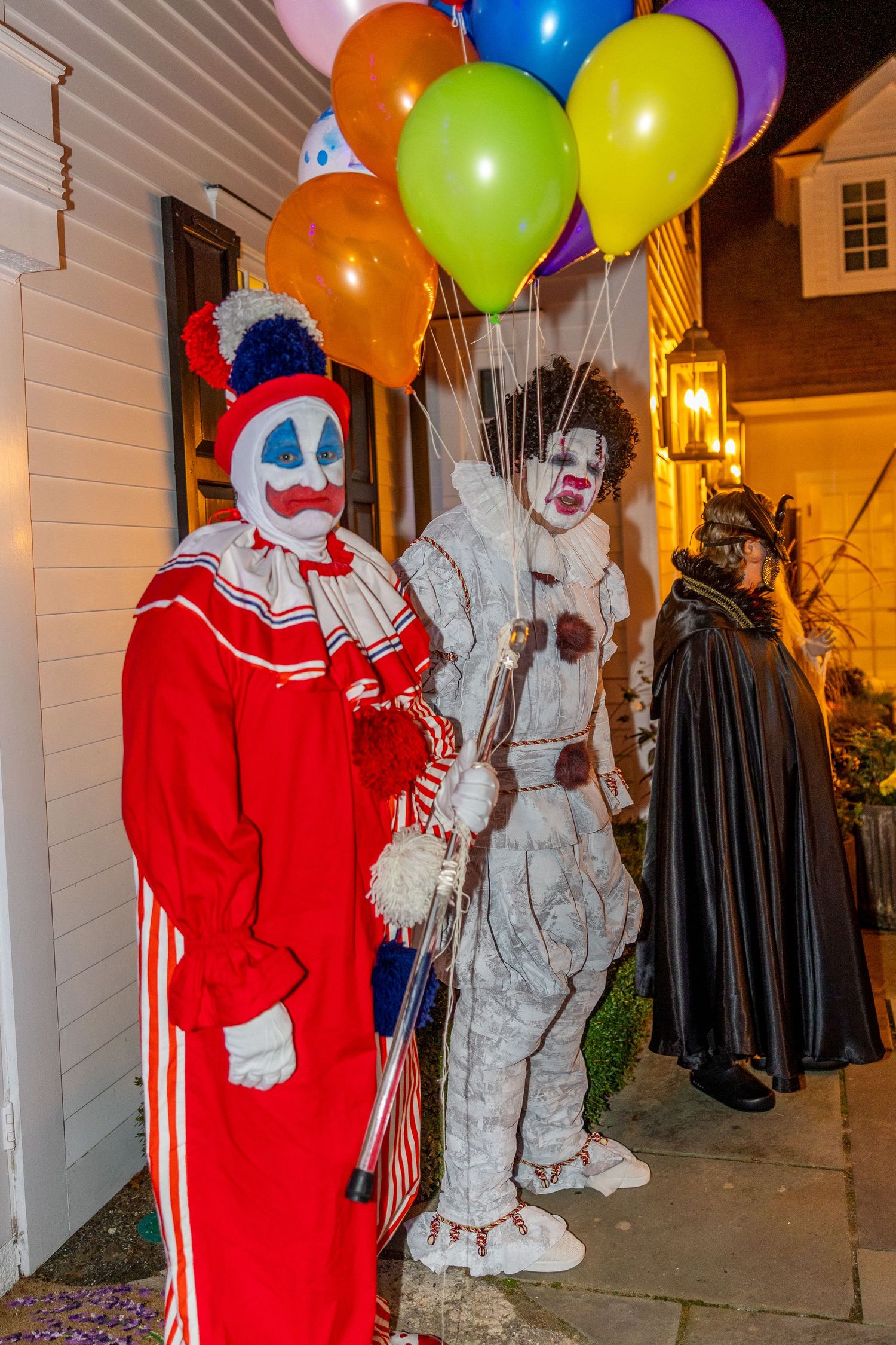 Three costumed figures including clowns with colorful balloons stand by doorway.