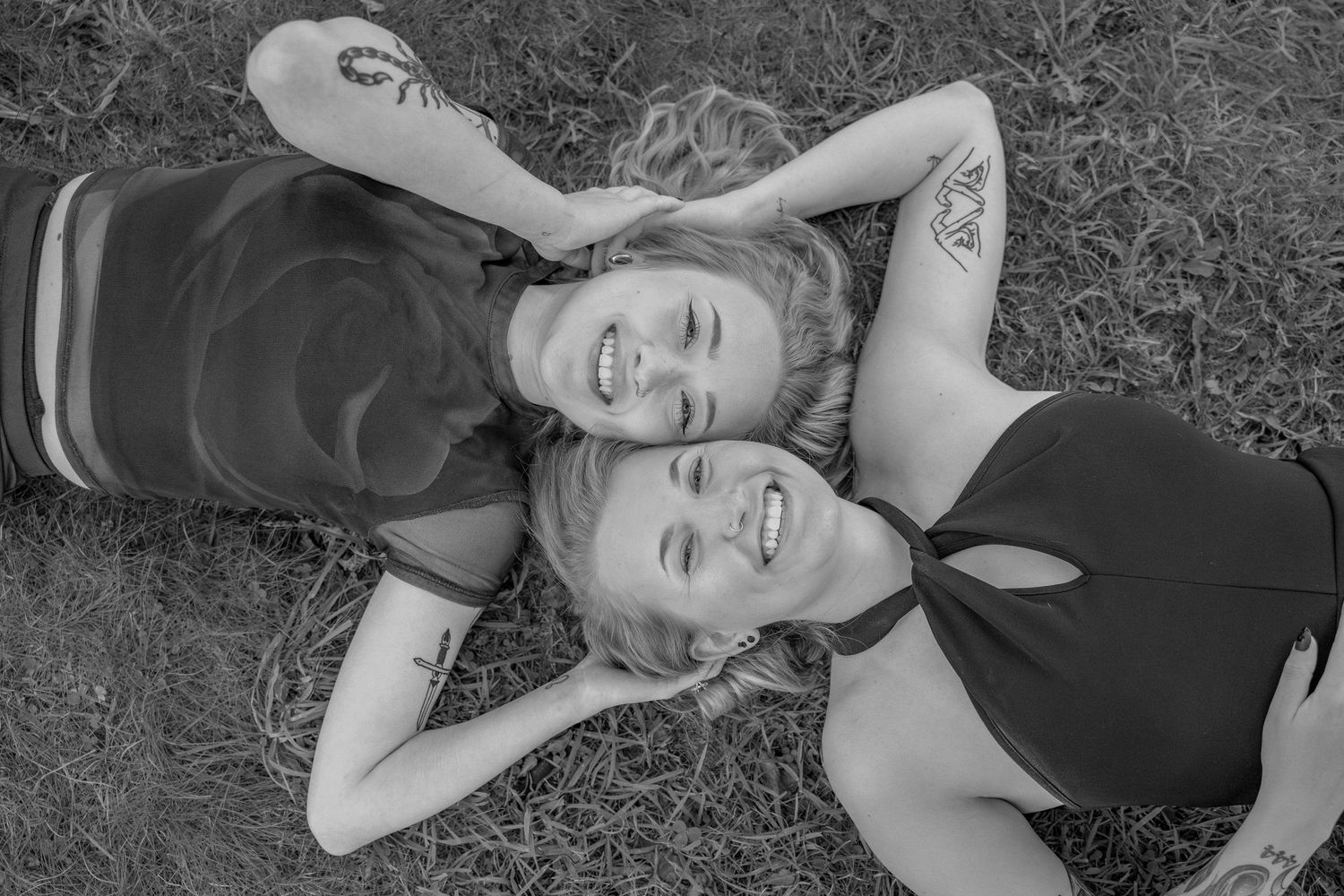 A black and white portrait of two people lying head to head on grass, sharing laughter.