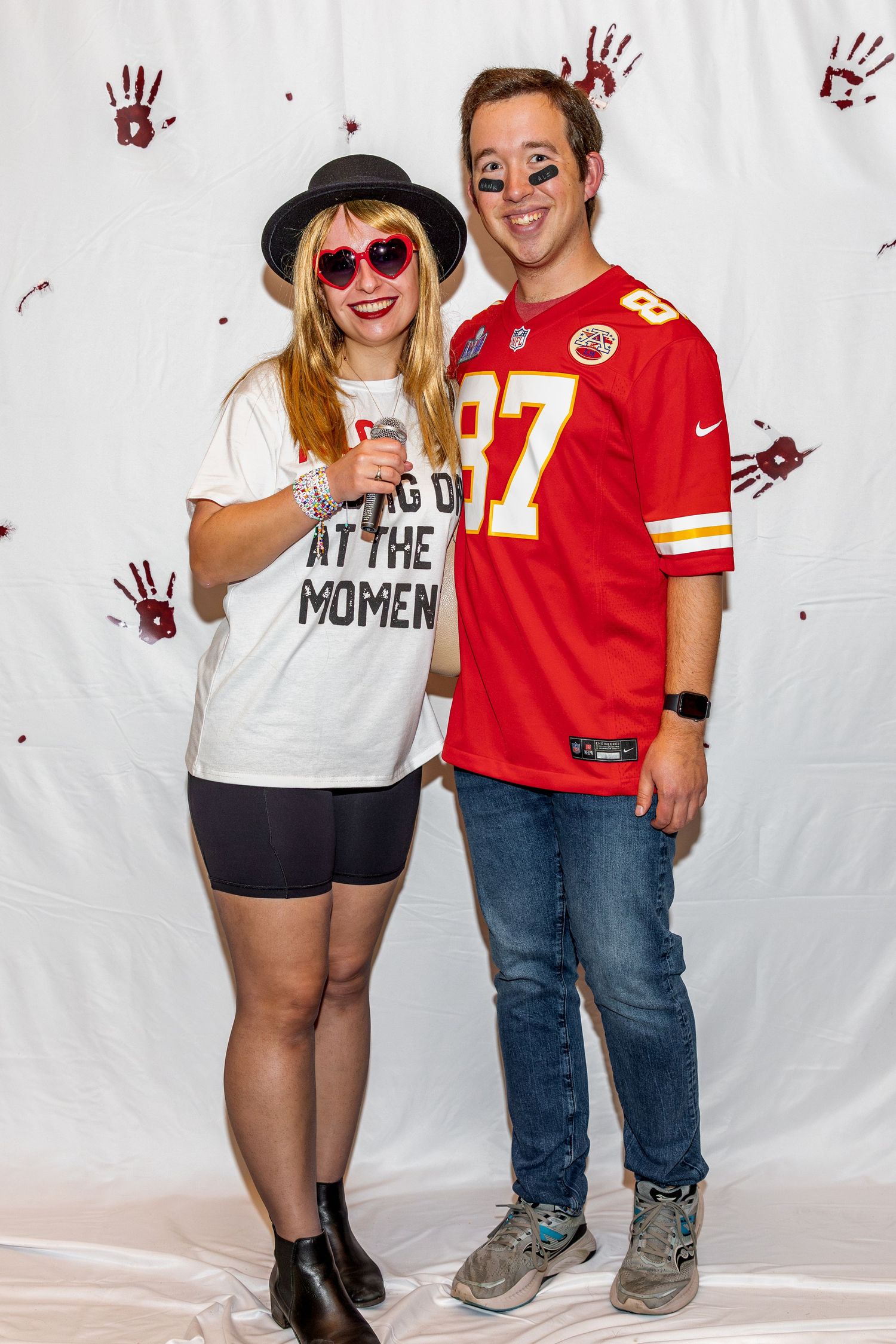 Two sports fans pose against white backdrop with red handprints wearing Chiefs jersey and casual attire.