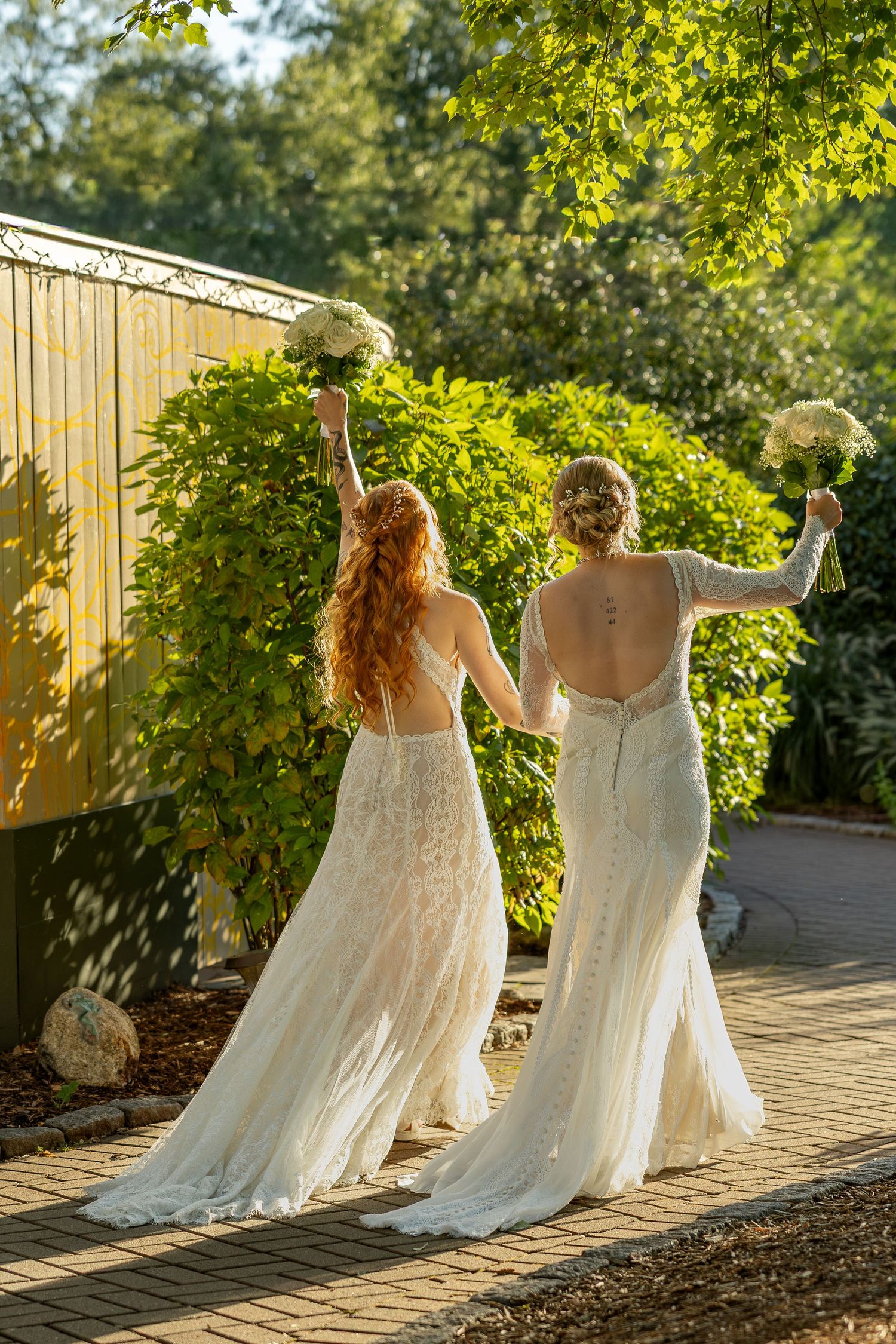 Two people in elegant white wedding dresses walking together on a garden path in golden sunlight.