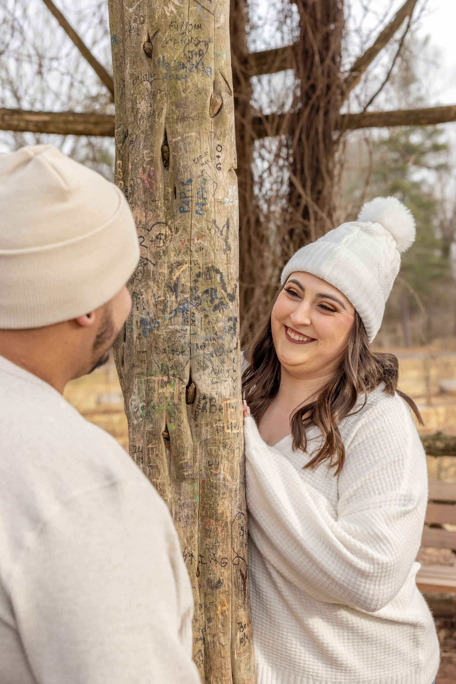 A playful winter engagement photo of a couple in white sweaters and knit beanies by a wooden post.