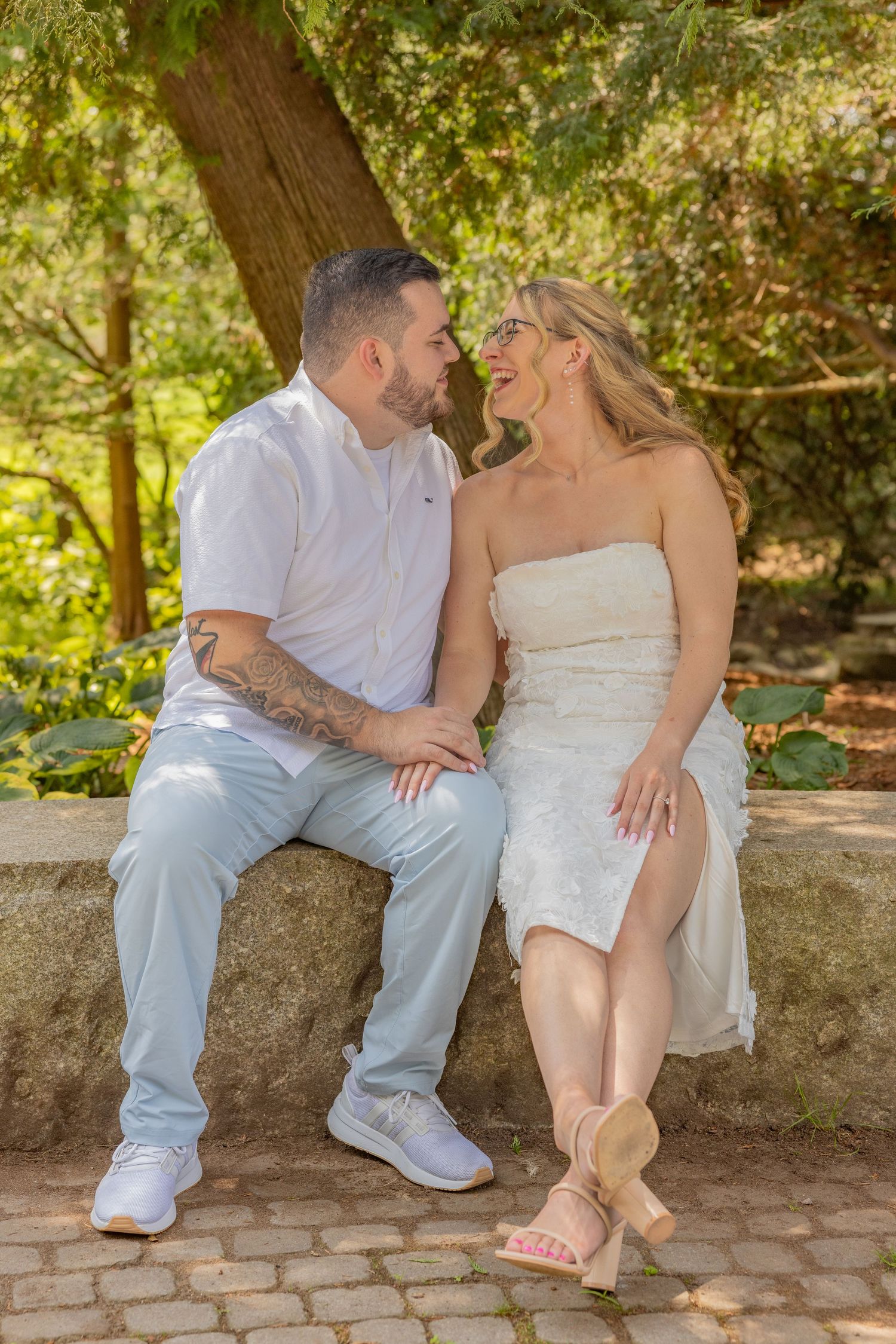 A couple shares a joyful moment while sitting on a stone wall in a garden setting.