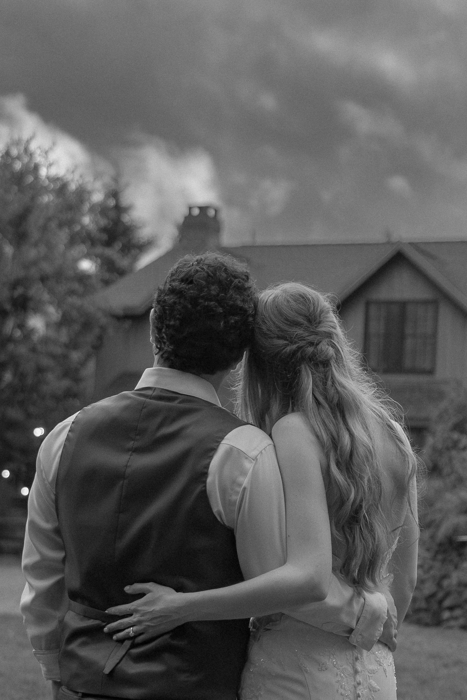 A couple in vintage clothing embraces while walking away from the camera in a black and white romantic scene.