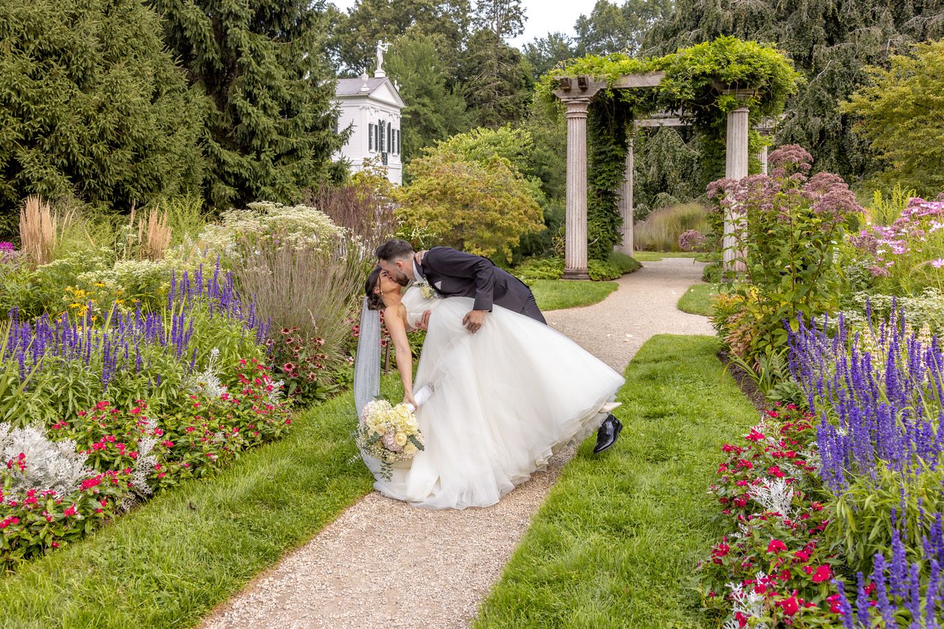 Wedding couple sharing romantic moment in lush garden with purple flowers and white pergola.