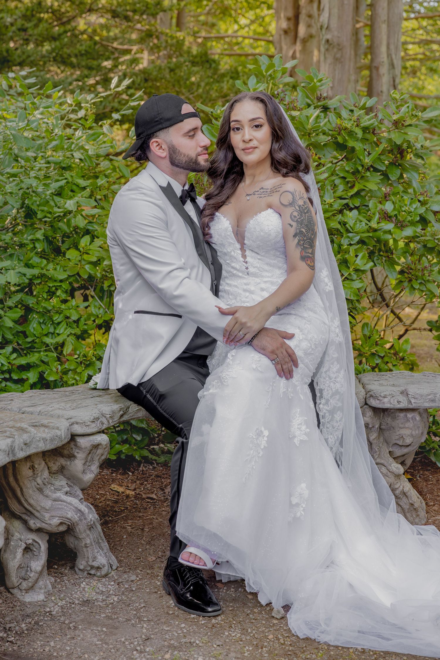 Bride in lace gown and groom in light suit sit together on stone bench in garden setting.