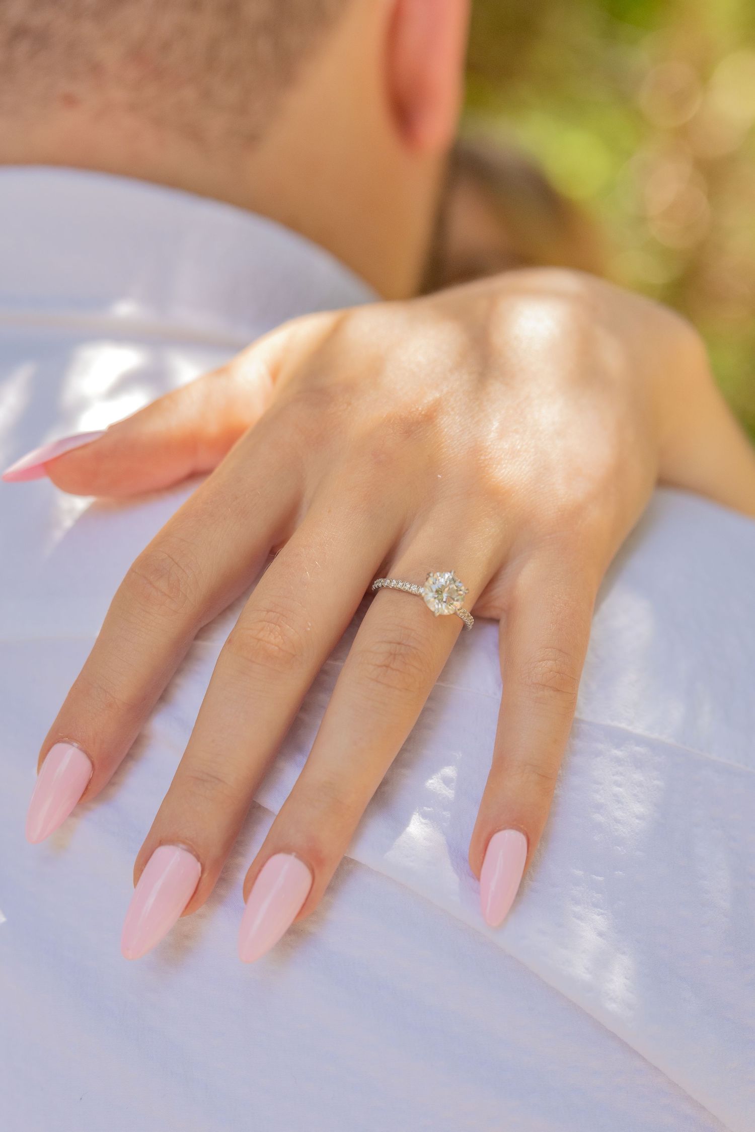 A perfectly manicured hand with pink nails displays a sparkling engagement ring against a white backdrop.