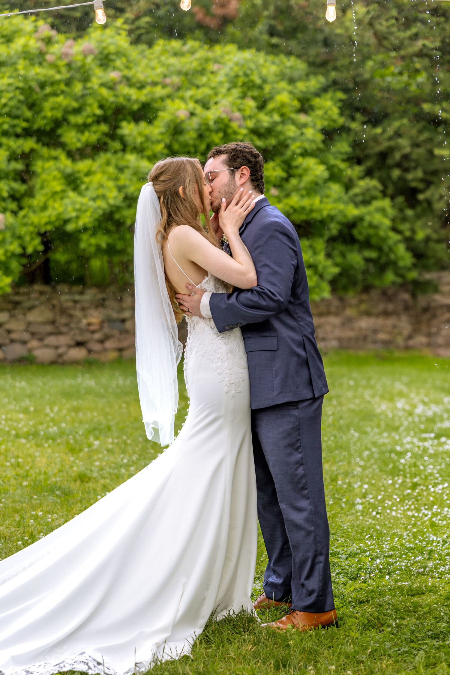 A romantic wedding day celebration showing a couple sharing a kiss on a lush green lawn surrounded by foliage.
