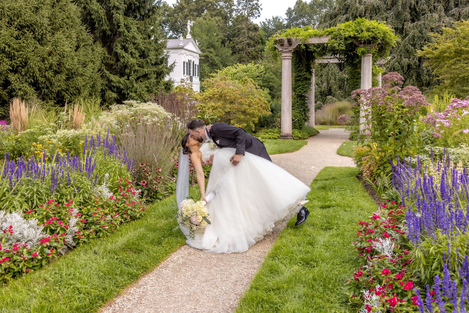 Wedding couple posing in garden with purple flowers and white pergola on sunny day.
