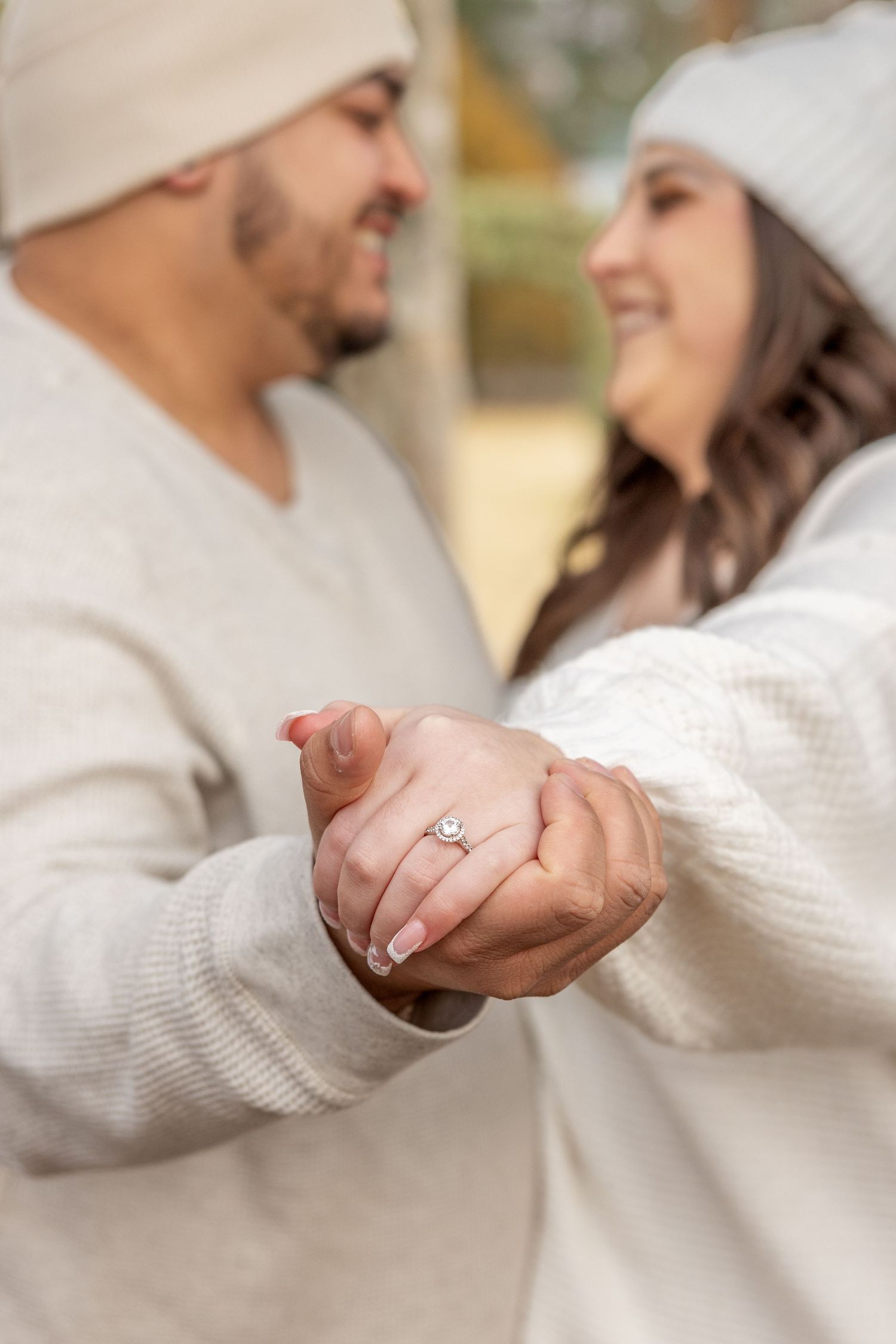 Engaged couple in cream sweaters and beanies laugh together while showing off the engagement ring.