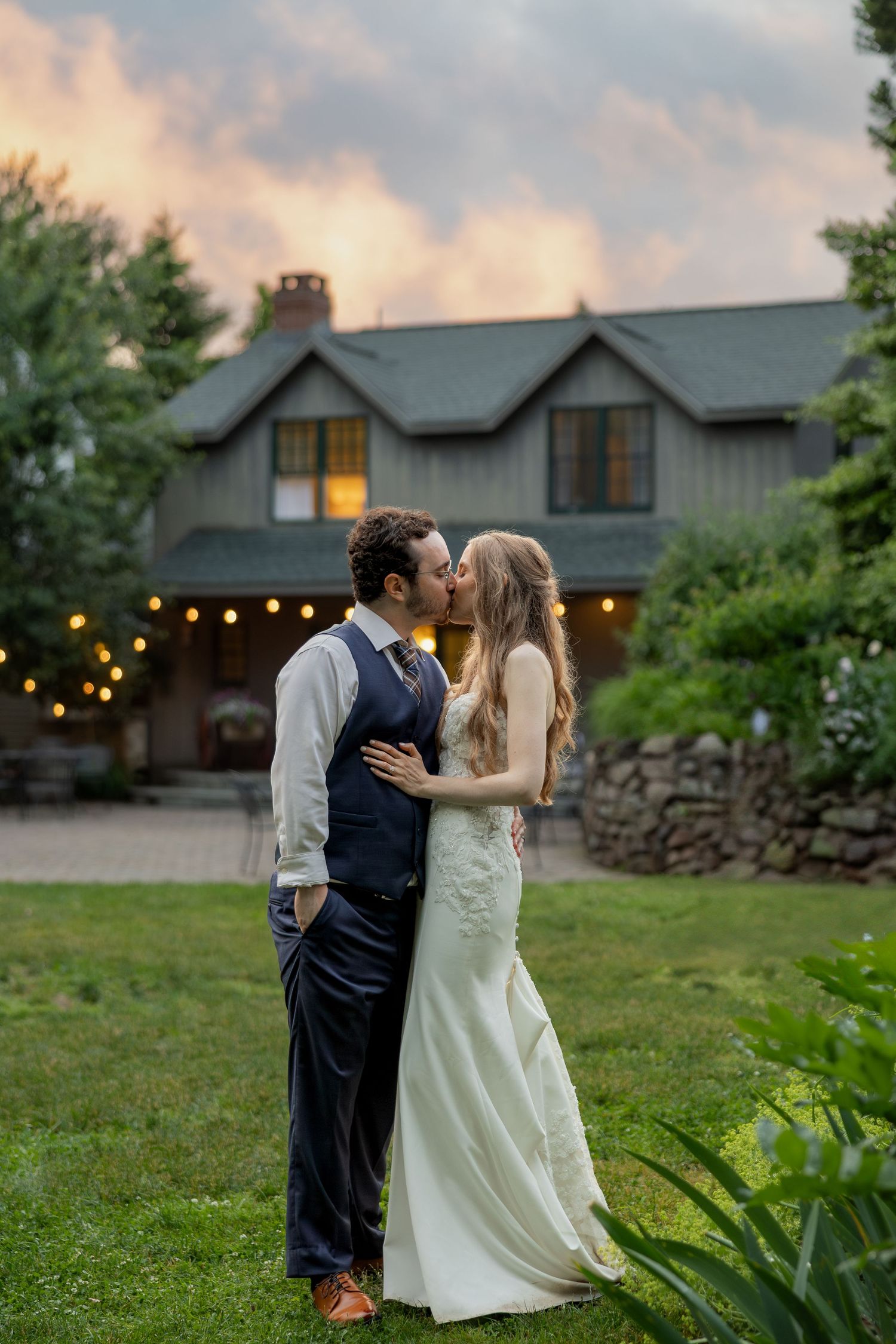 A couple shares a romantic kiss at dusk outside a rustic house with string lights and a beautiful garden setting.
