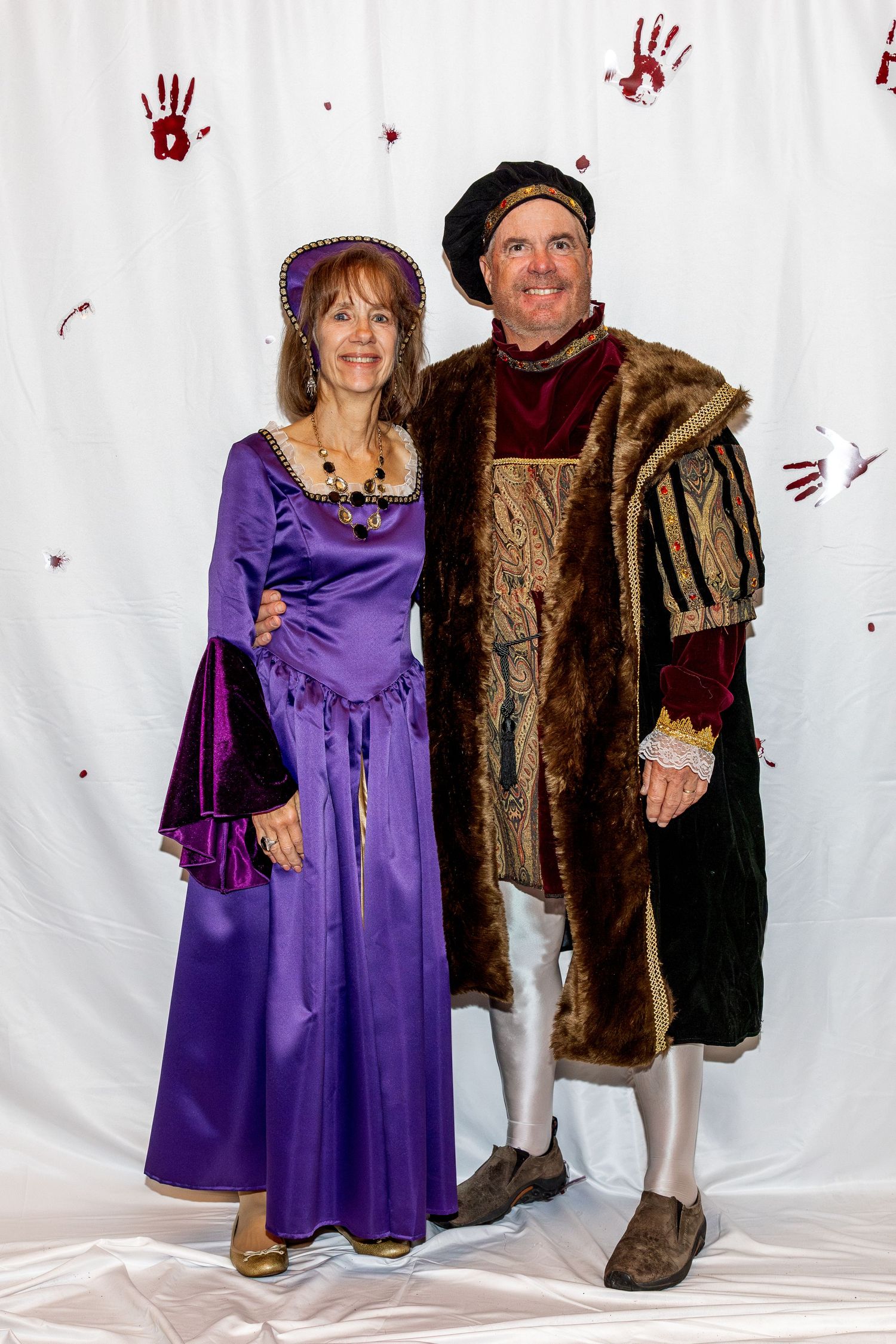 Couple in medieval renaissance festival costumes posing against white backdrop with red handprint decorations.