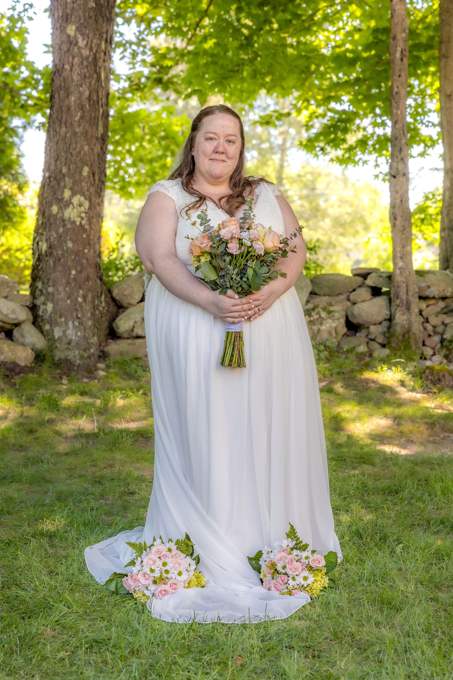 A bride in a flowing white dress poses with her bouquet in front of a rustic stone wall and trees.