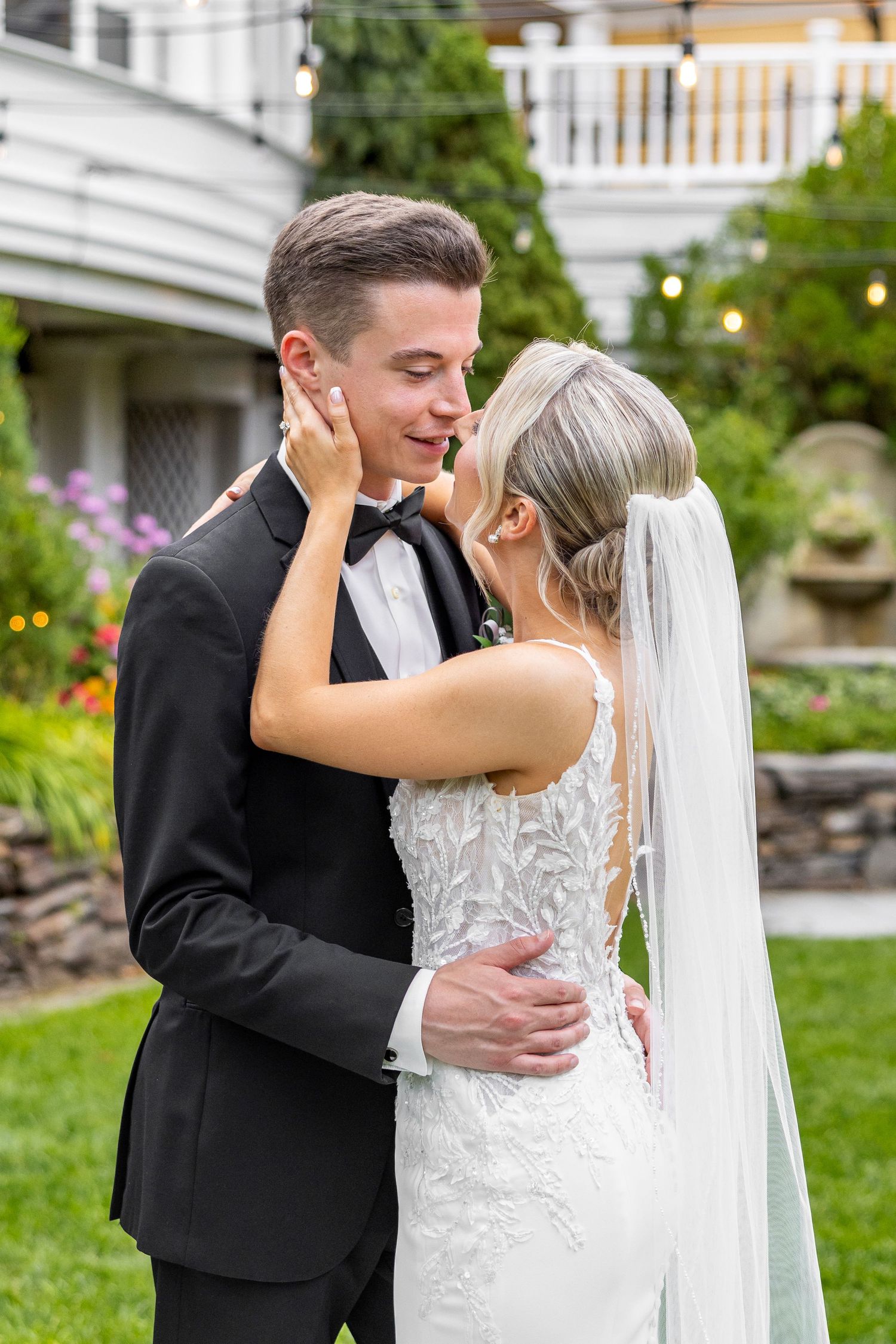 Tender moment between bride and groom in garden setting with white house and flowers in background.