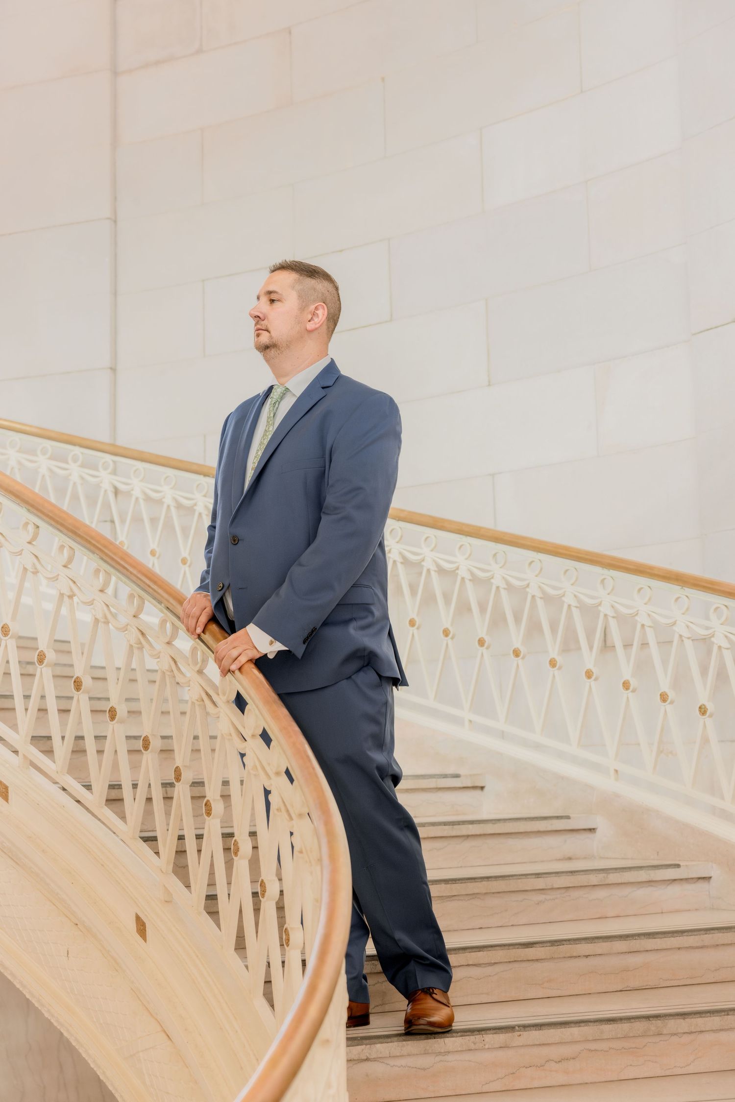 Groom in navy suit stands on elegant curved marble staircase.