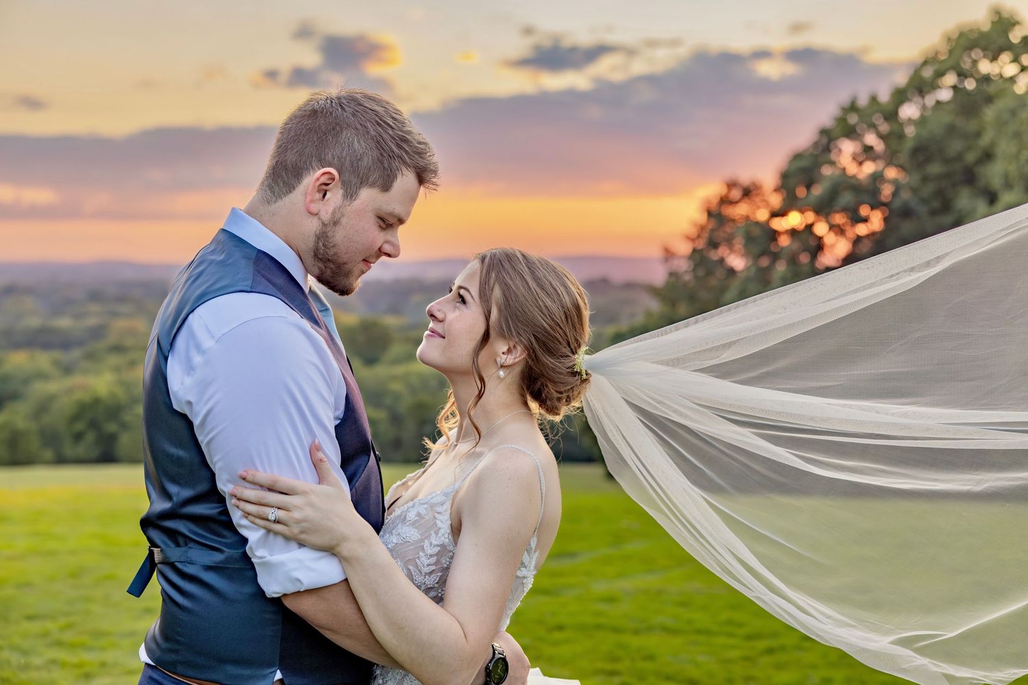 Intimate wedding portrait at sunset with flowing veil and rolling hills in background.