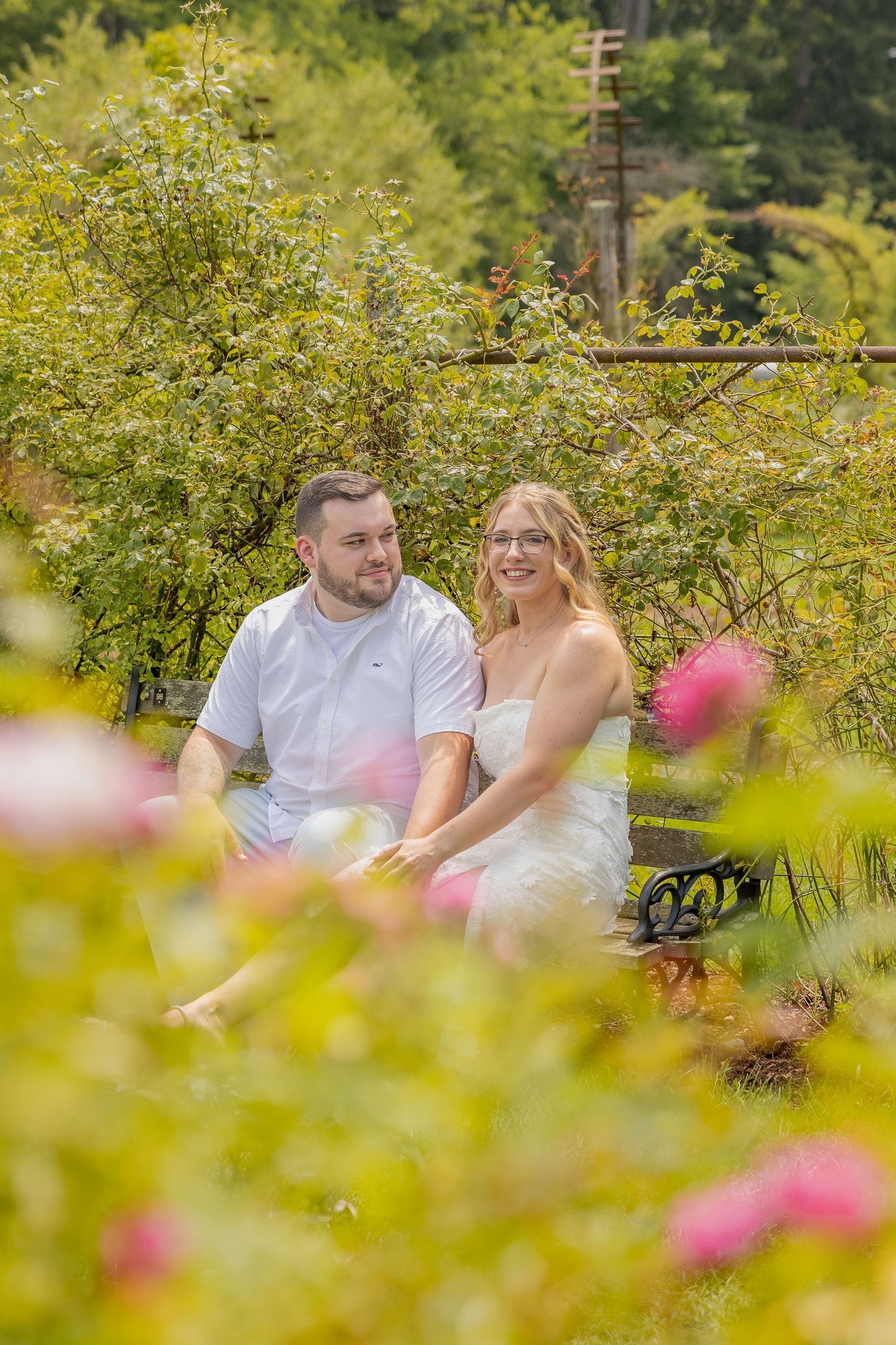 A couple sits together on a garden bench surrounded by lush greenery and pink flowers on a sunny day.