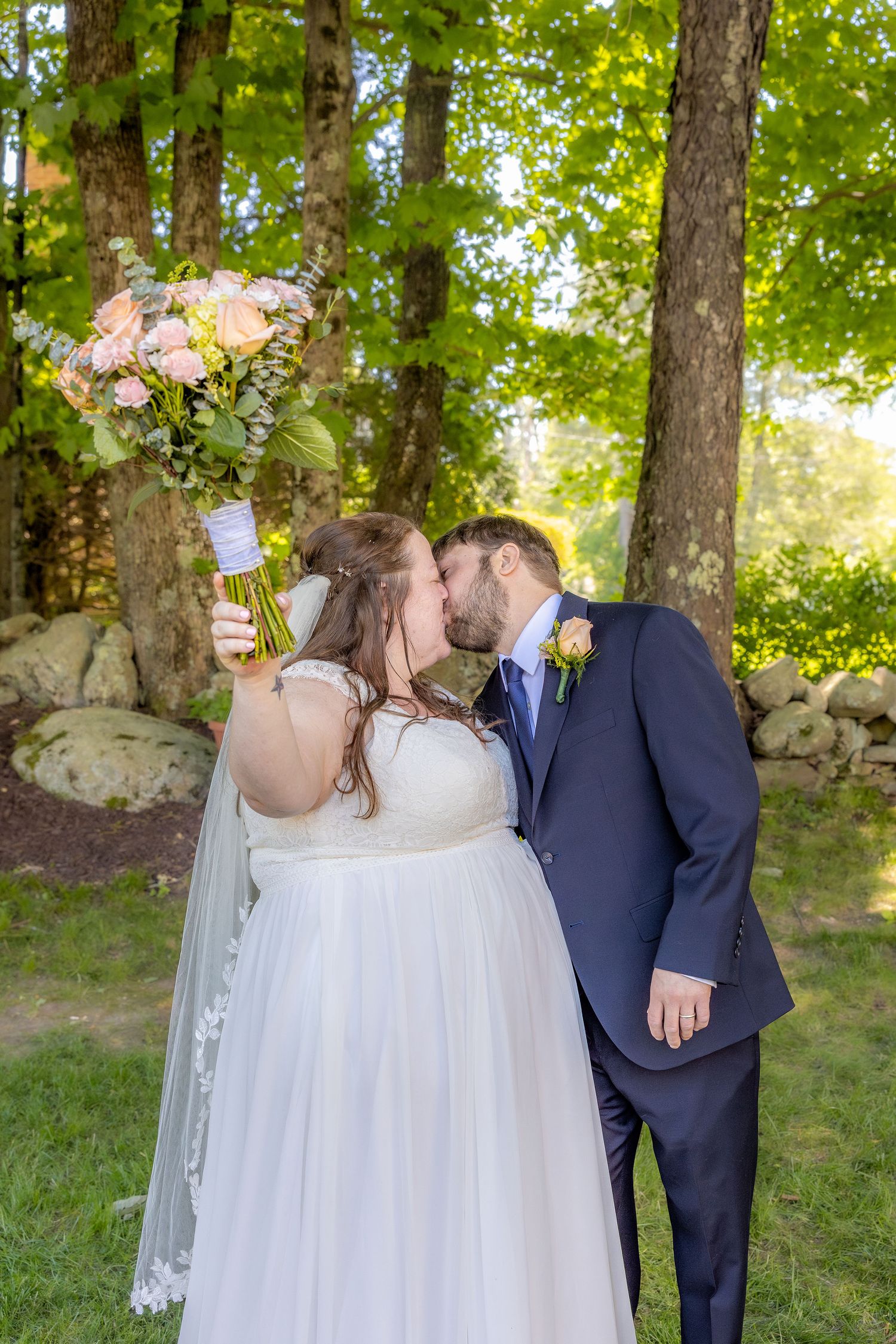 A couple shares their first kiss during their outdoor wedding ceremony against a backdrop of trees and stone wall.