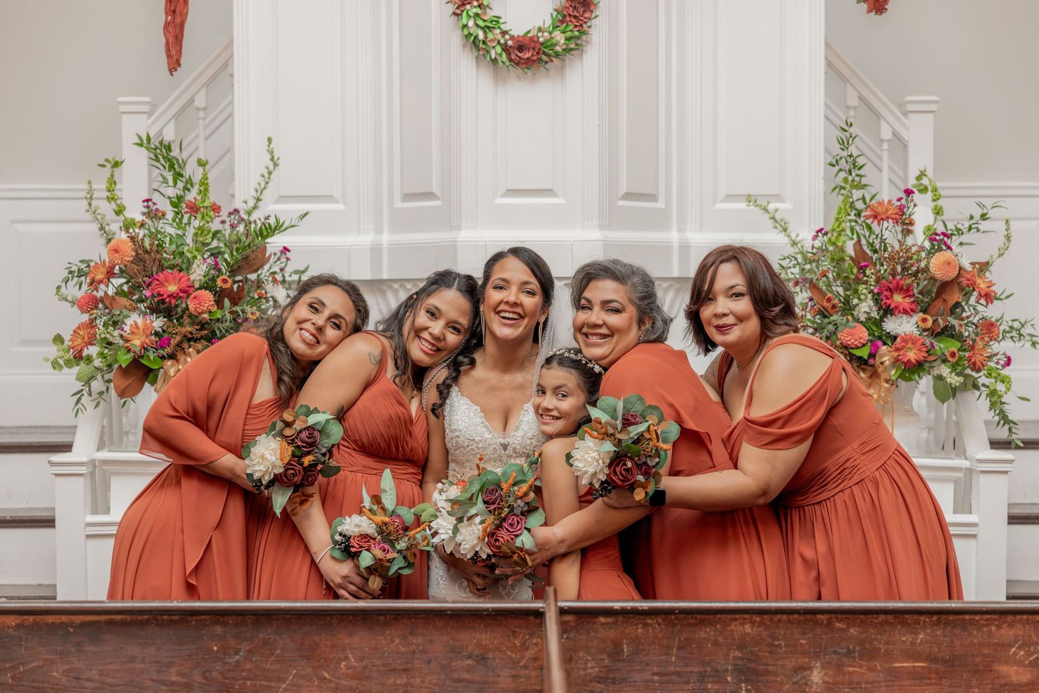 Bridal party in coral dresses poses joyfully on church steps with floral arrangements.