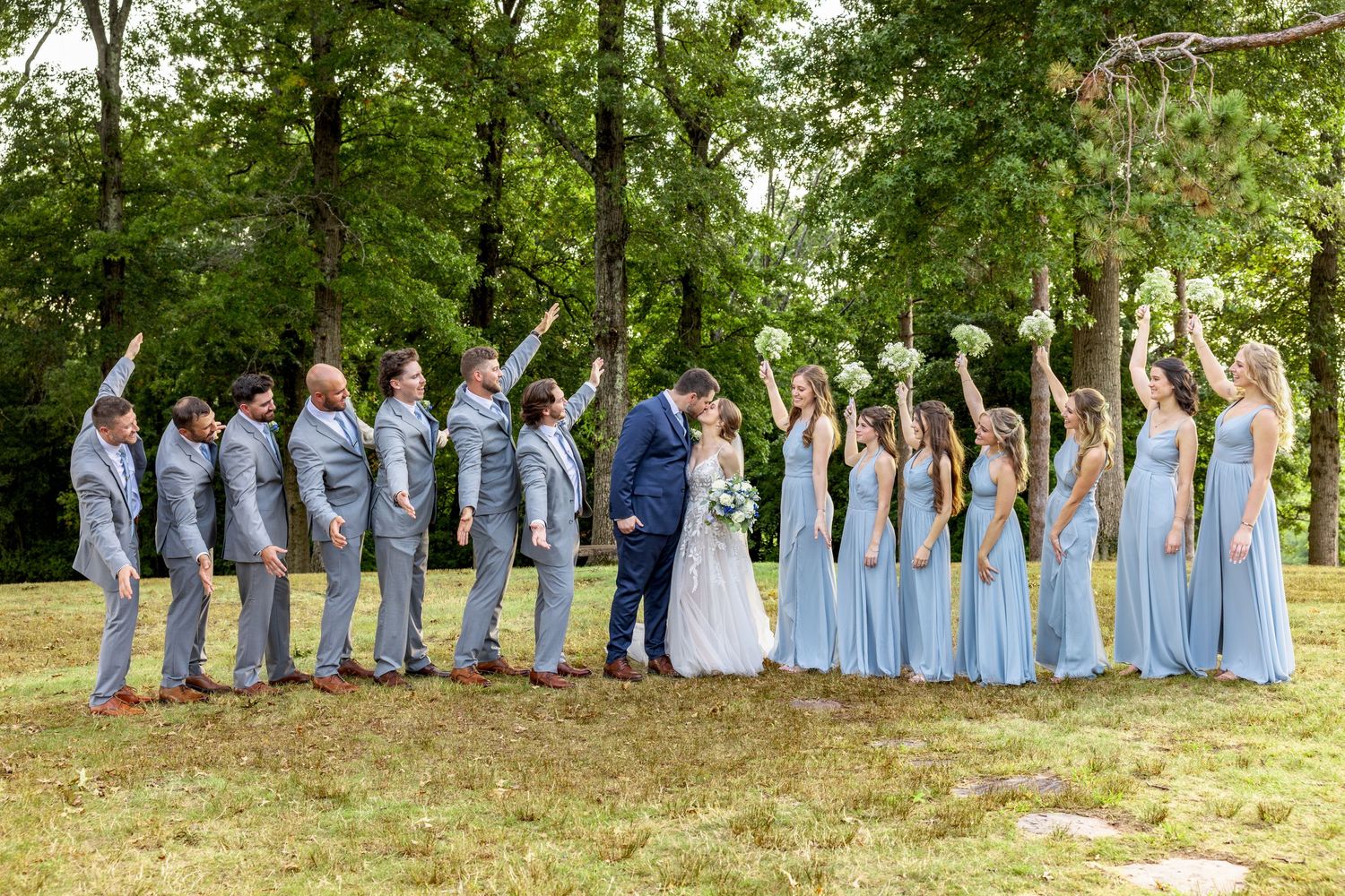 Wedding party lined up outdoors wearing light blue dresses and grey suits under green trees.