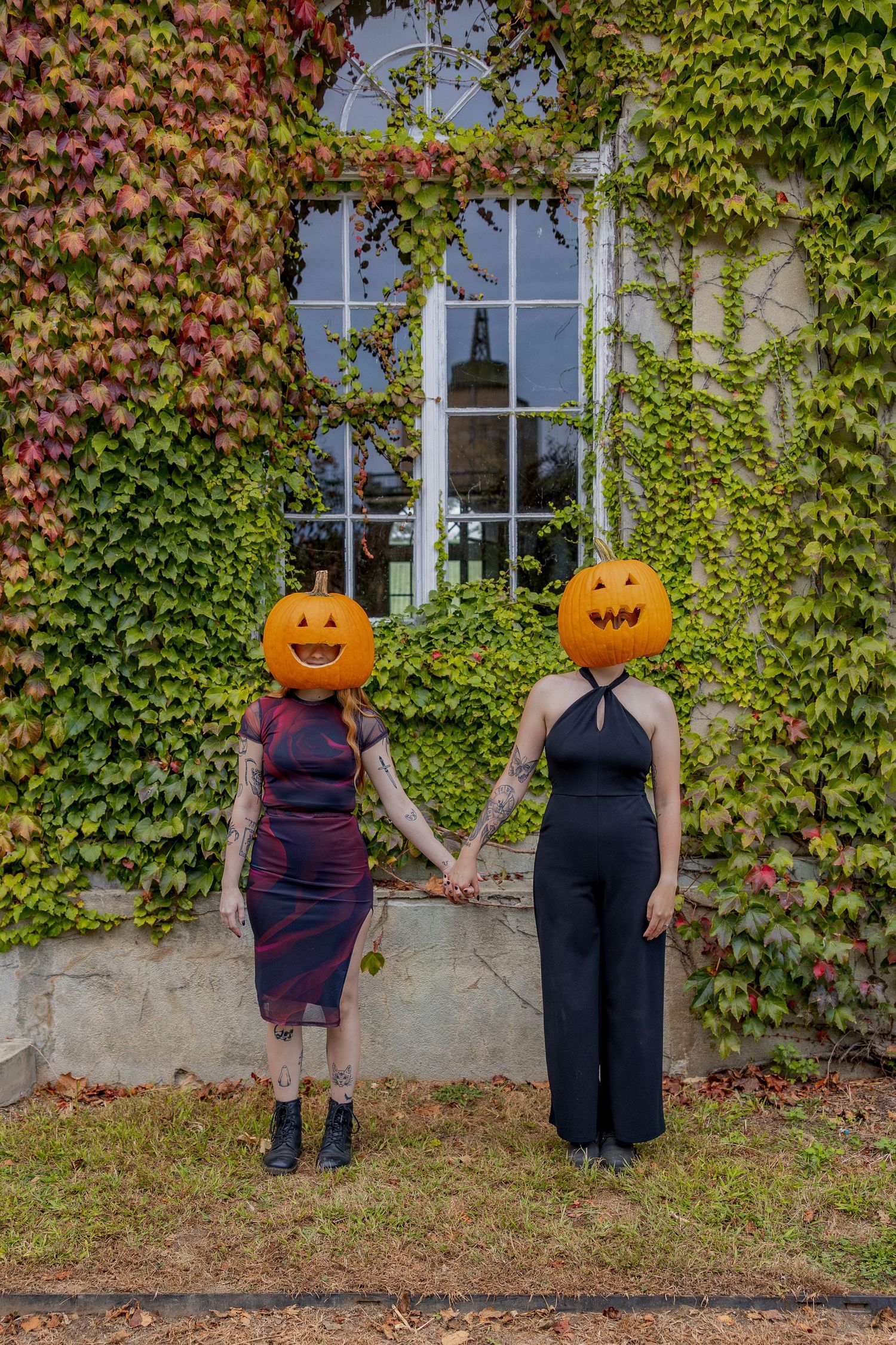 Two people in formal attire wearing pumpkin heads hold hands in front of an ivy-covered wall on Halloween.